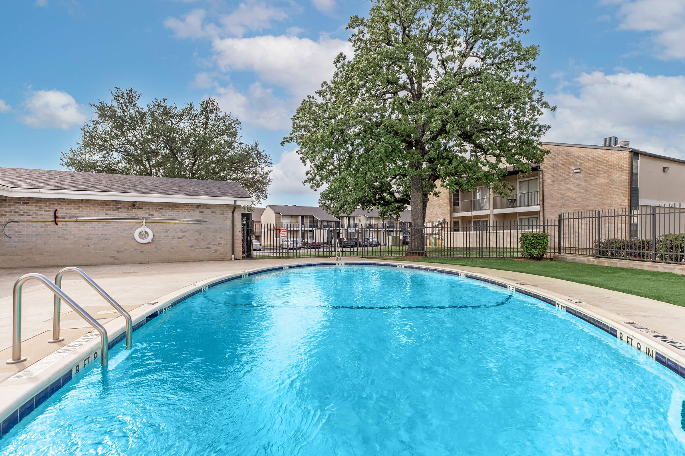 A clear blue swimming pool surrounded by a fenced area, with a tree nearby and apartment buildings in the background. The sky is partly cloudy, creating a bright and inviting atmosphere for relaxation and enjoyment.