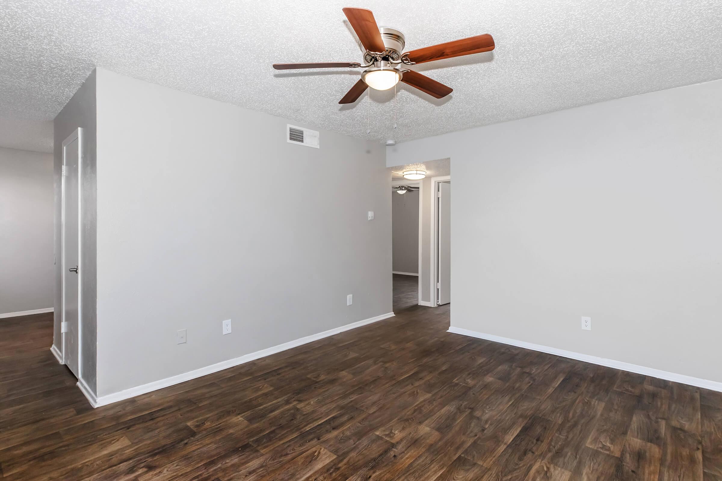A spacious, well-lit living room featuring light gray walls and dark hardwood floors. A ceiling fan with wooden blades is mounted on the ceiling. The room leads to a doorway, indicating access to additional areas. Natural light adds warmth to the space.