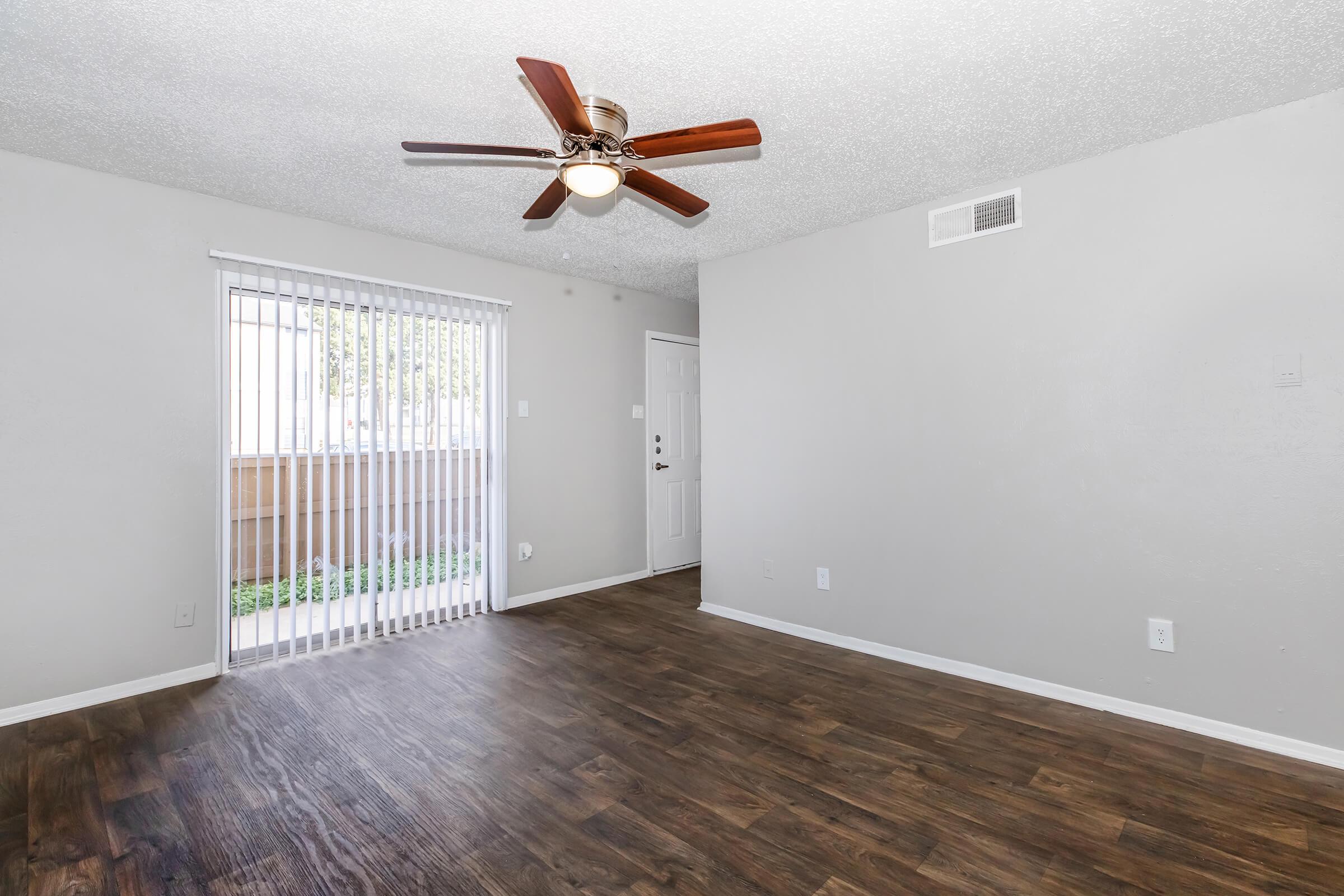 A spacious room featuring a ceiling fan, light wood-look flooring, and neutral gray walls. There is a sliding glass door with vertical blinds leading to an outdoor area, and a front door visible on the right side. The room is empty, providing a blank canvas for interior decorating.