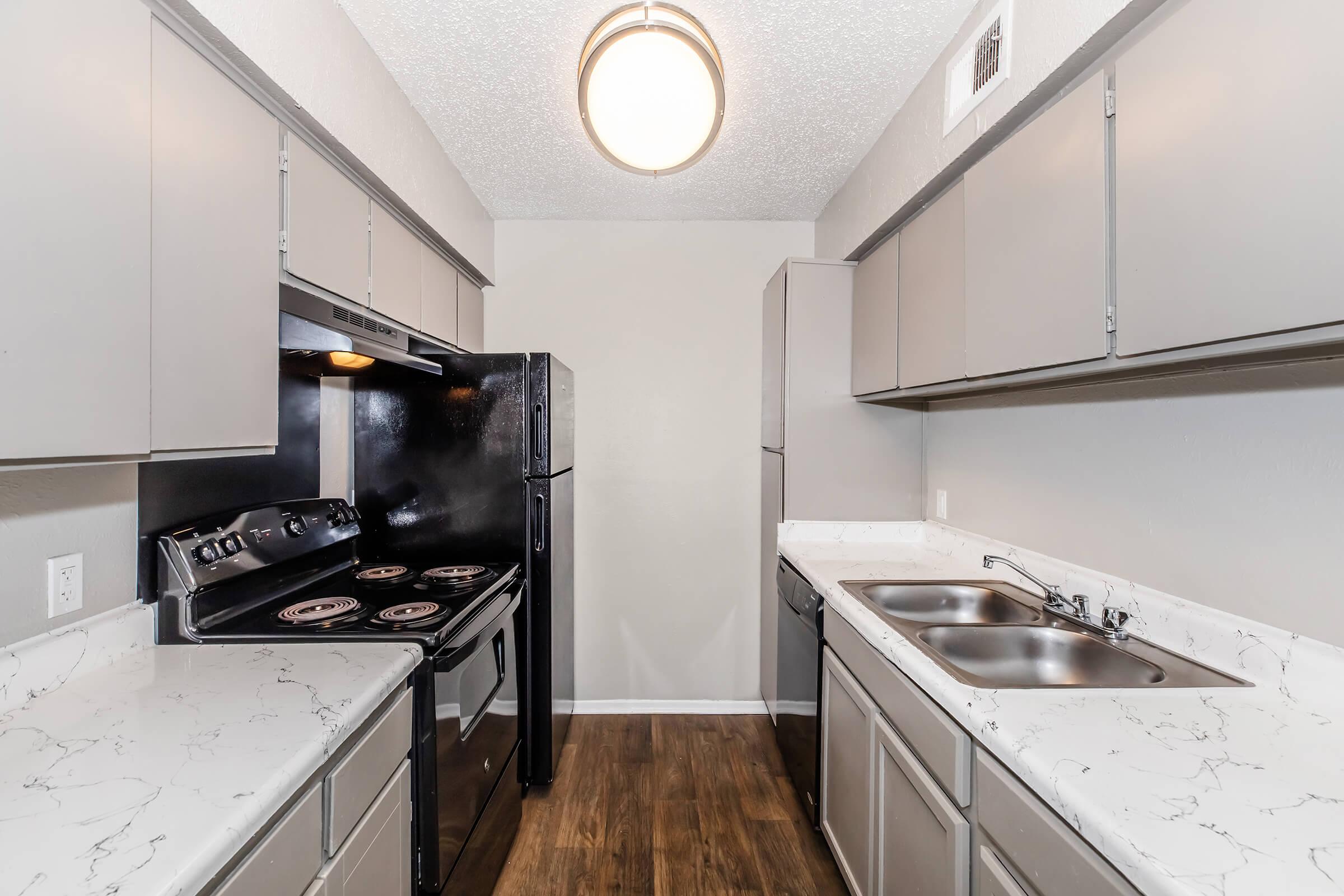 Modern kitchen featuring gray cabinets, a black stove and refrigerator, white countertops, and dual sinks. The space has wood-style flooring and a ceiling light fixture, creating a clean and contemporary look.