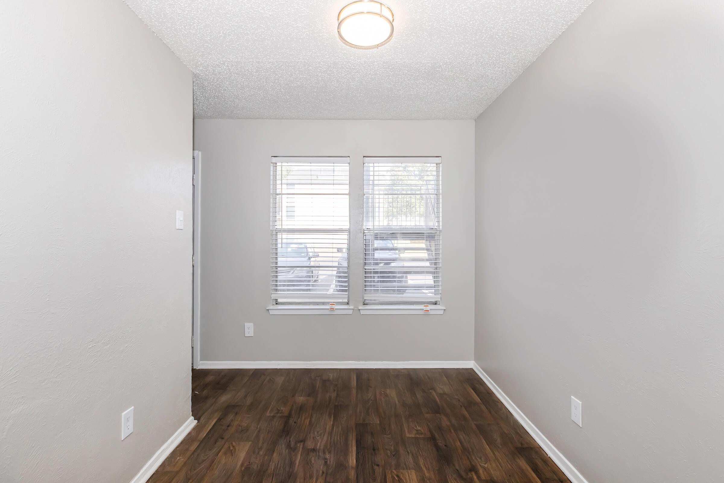 Empty room with light gray walls and wooden flooring. There are two windows with blinds allowing natural light, and a round ceiling light fixture. The space is uncluttered and ready for furniture arrangement.