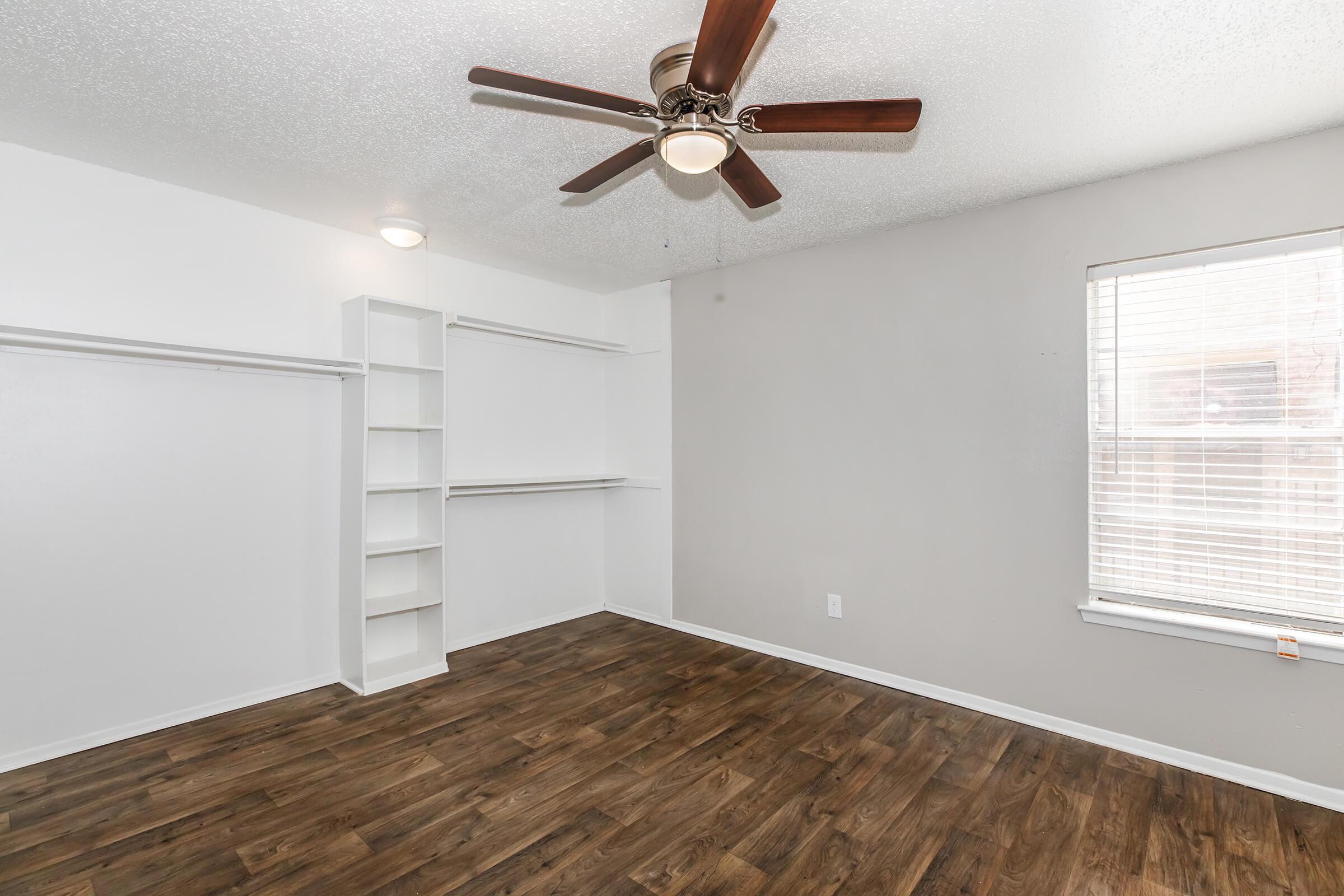 A bright, spacious room with light-colored walls and a ceiling fan. It features a window with blinds, hardwood-style flooring, and built-in shelving along one wall next to a closet. The overall atmosphere is clean and inviting, offering plenty of natural light.