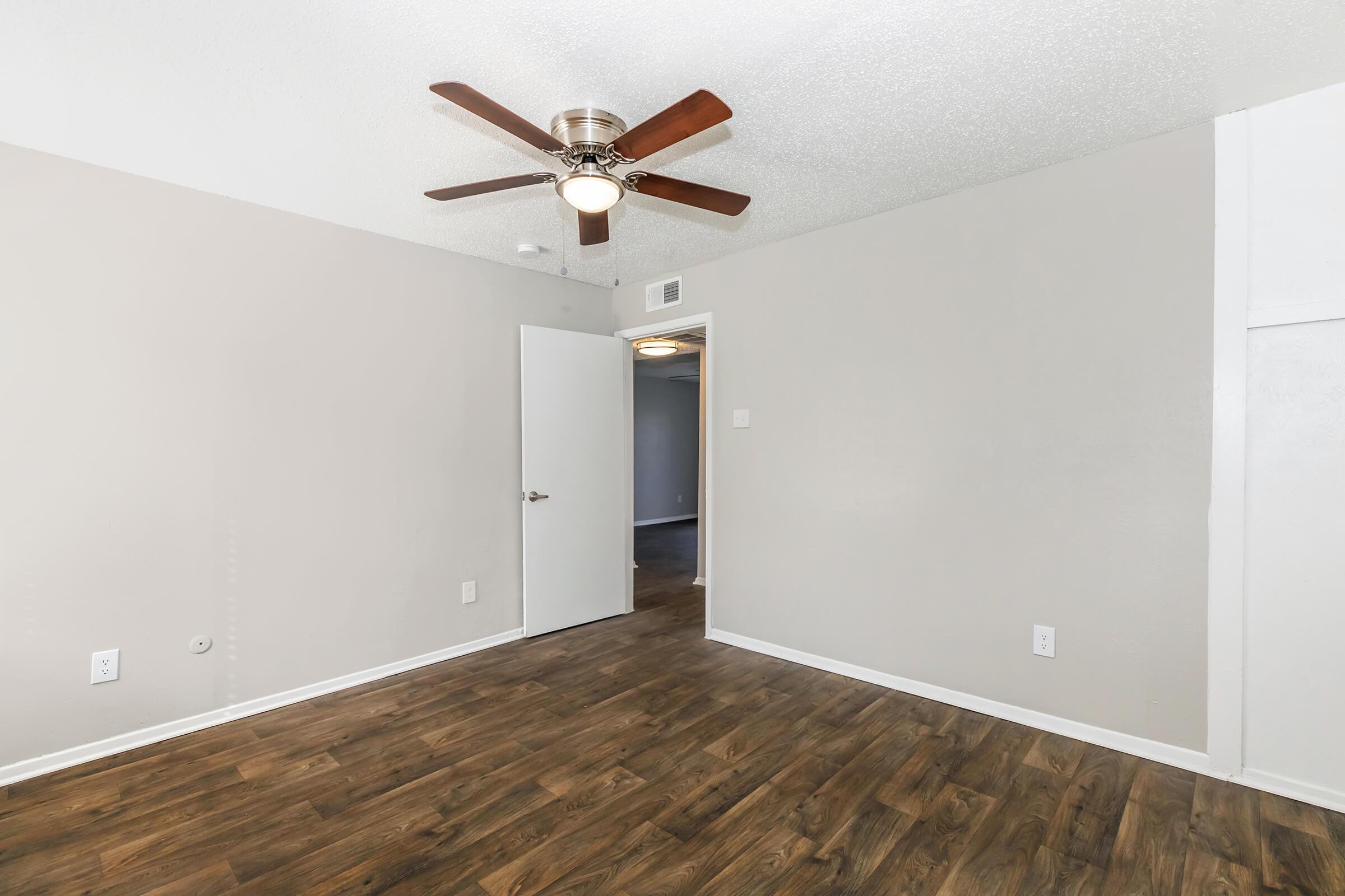 A well-lit room featuring a ceiling fan and light fixture, with light gray walls and dark wood floors. An open doorway leads to another room, while the opposite wall remains blank. The room appears spacious and inviting, suitable for various furniture arrangements.