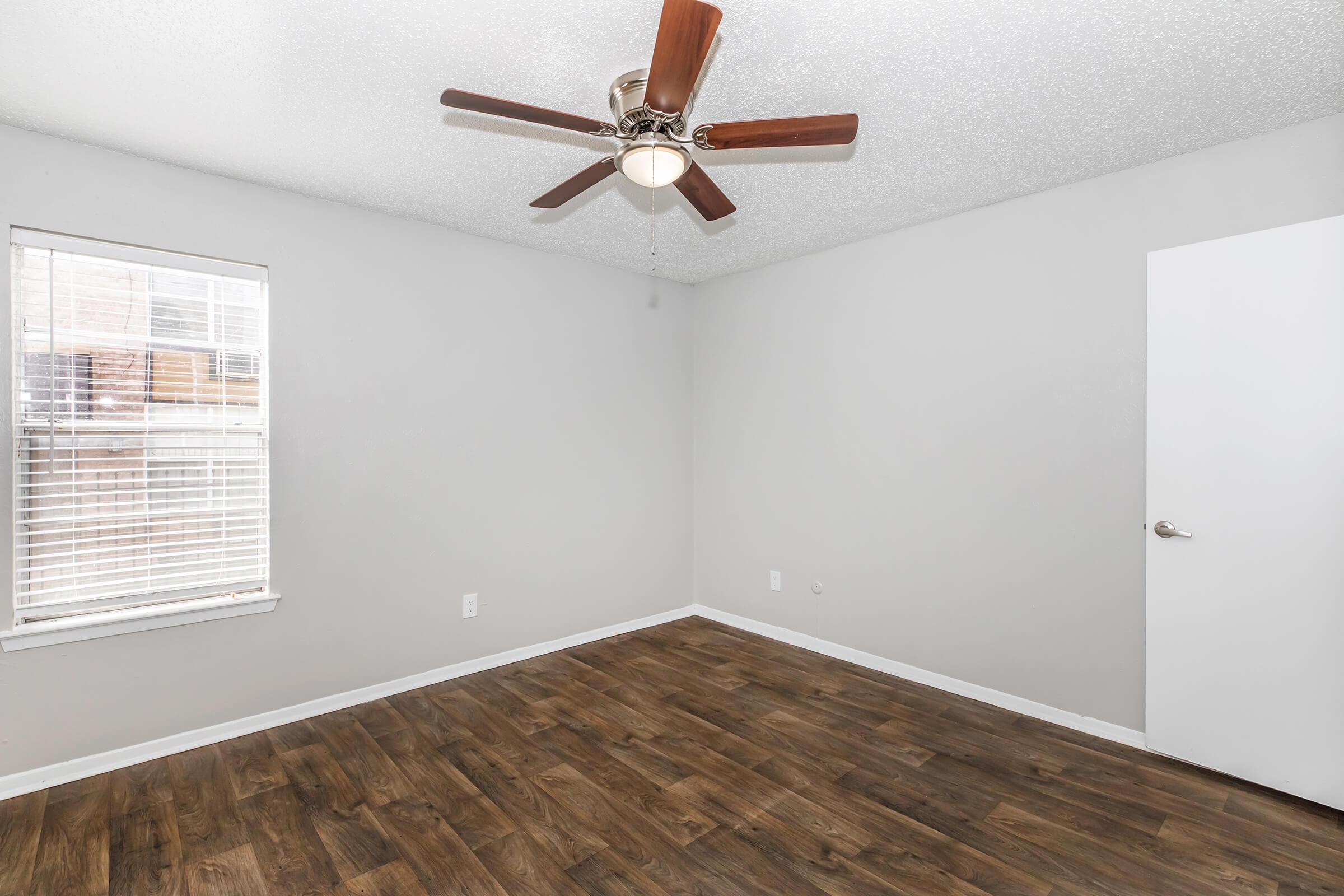 A bright, empty room with light gray walls and a ceiling fan. The floor is covered in dark wood laminate. There's a window with blinds letting in natural light, and a door on the right side. The space is open and ready for furnishing.