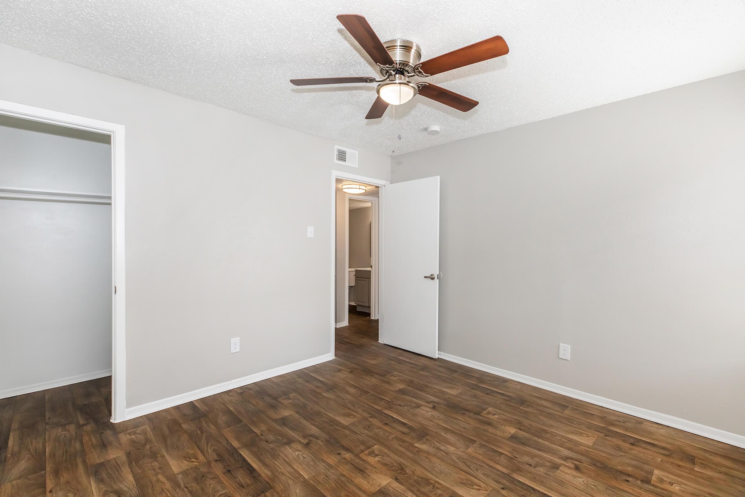 A well-lit room with a wooden floor, featuring a ceiling fan with wooden blades. There are two doors: one leading to a closet and the other to a hallway. The walls are painted a soft gray, and the overall atmosphere is clean and modern.