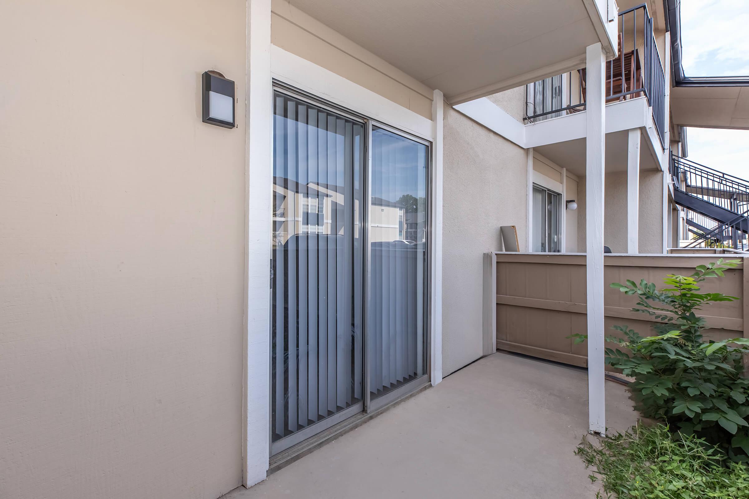A view of a residential patio area featuring sliding glass doors with vertical blinds, a concrete floor, and a beige wall. There is a small wooden fence and some greenery visible nearby, along with part of a multi-story building in the background.