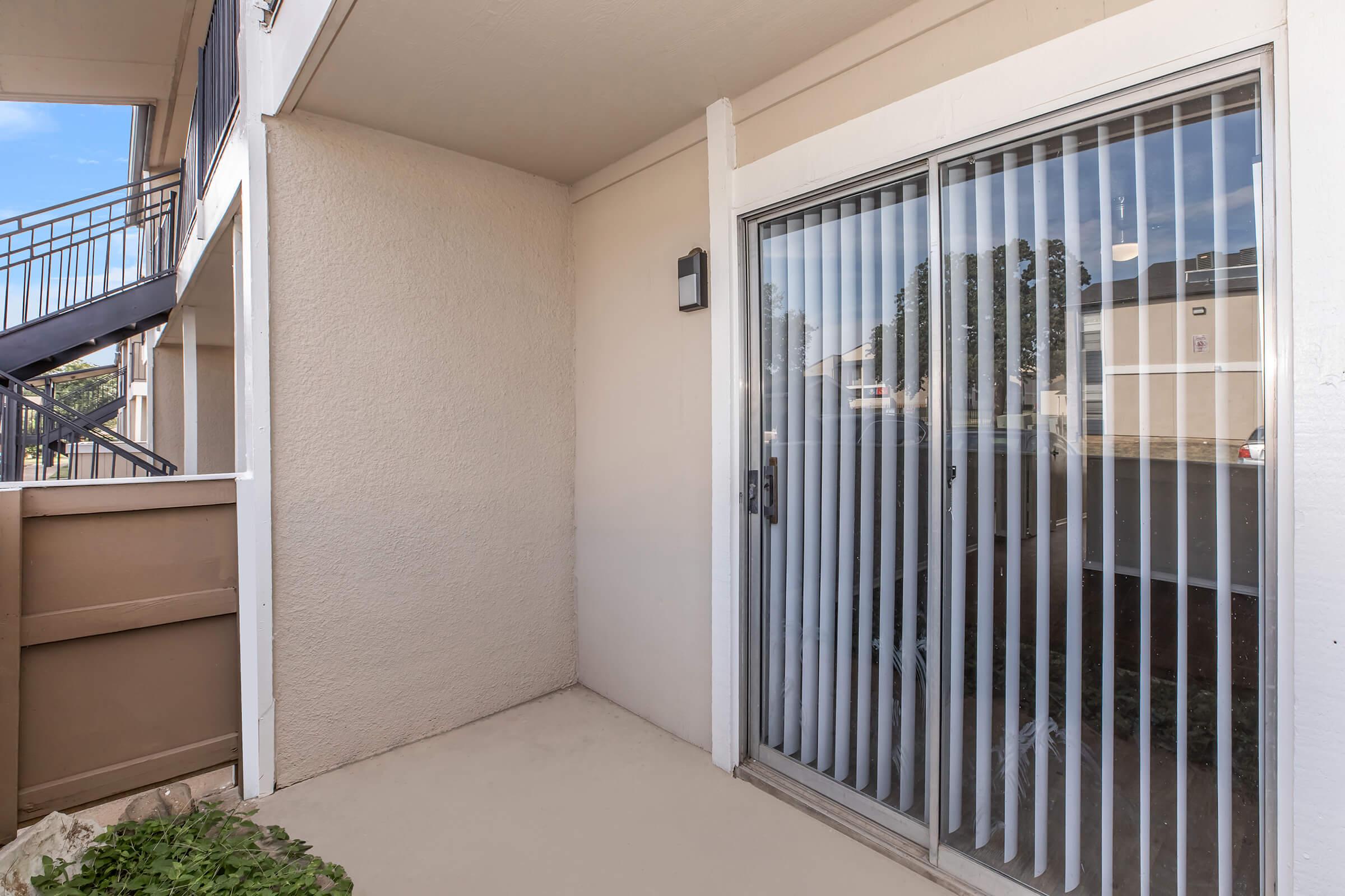A small, covered patio area with sliding glass doors featuring vertical blinds. The wall is a light beige color, and a light fixture is mounted beside the door. Thereโs a staircase visible in the background, leading up to another level of the building. The floor is plain and lacks furnishings.