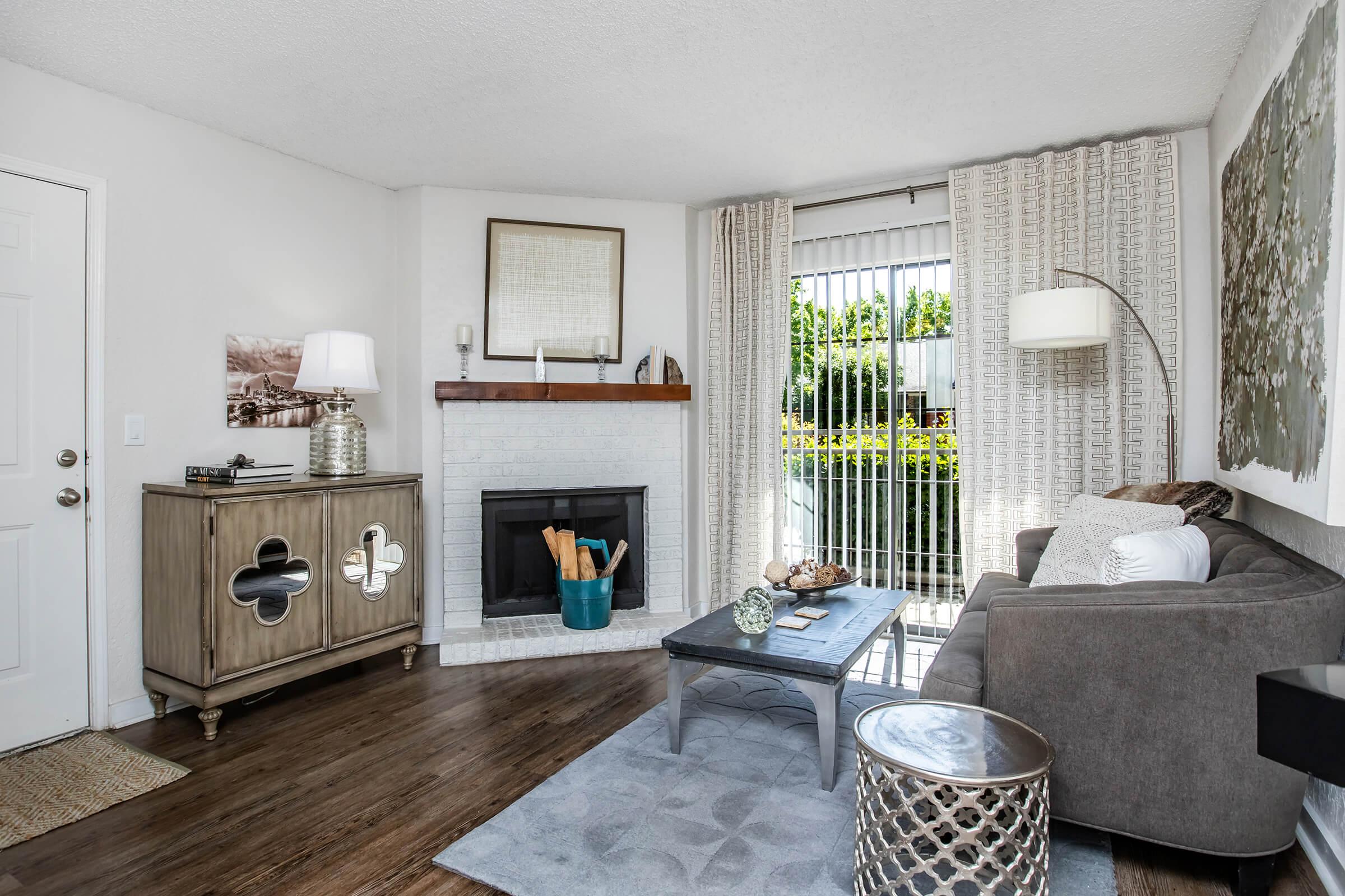 A cozy living room featuring a gray sofa, a wooden coffee table, and a decorative rug. There's a fireplace with a wooden mantel, a lamp on a console table, and large windows with white curtains letting in natural light. Greenery can be seen outside the window, adding a touch of nature to the space.