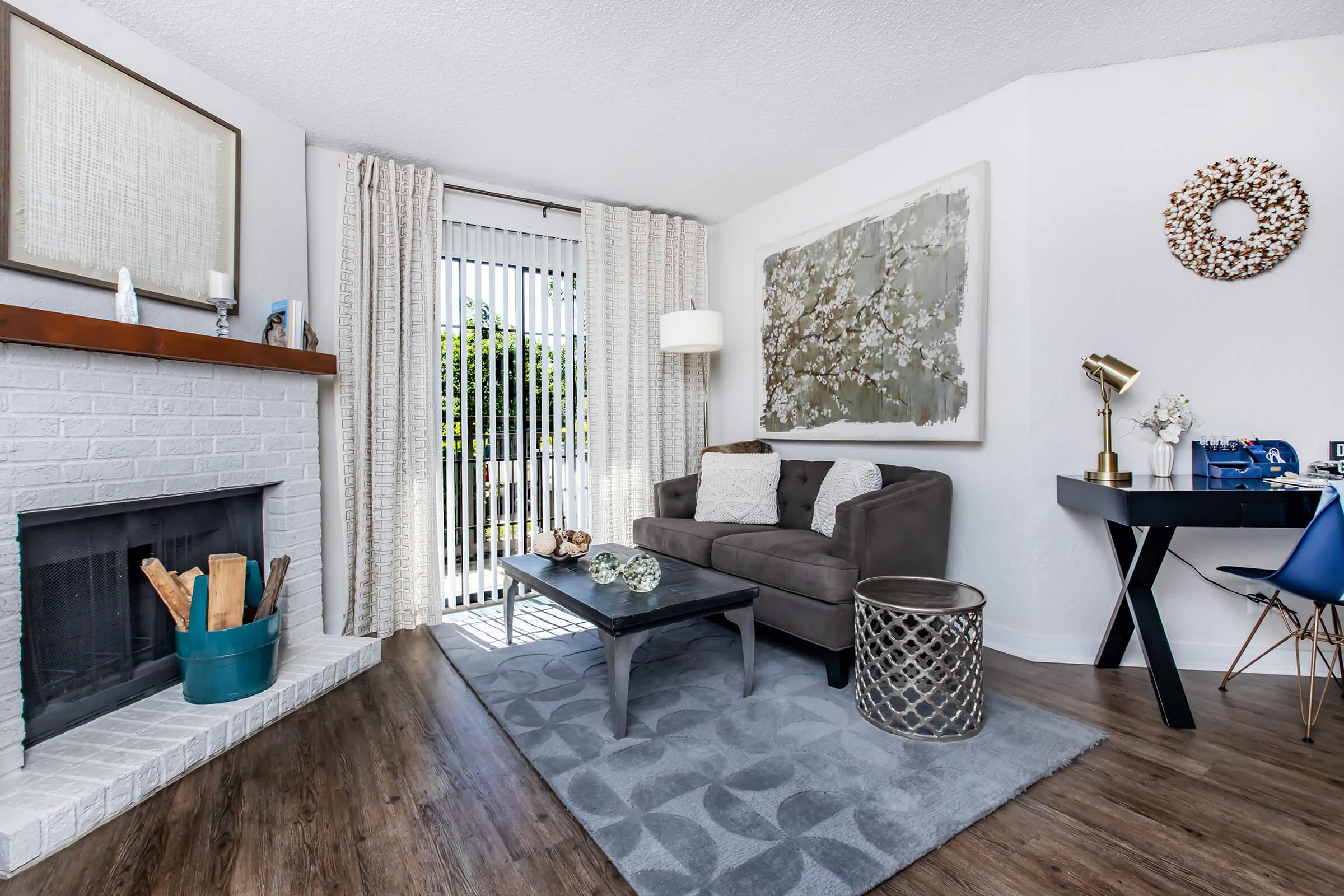 A cozy living room featuring a gray sofa, a glass coffee table, and a decorative area rug. There’s a fireplace with firewood, a small side table, and a desk with a blue chair. Soft curtains cover the sliding glass door, allowing natural light to brighten the space, complemented by artwork on the walls.