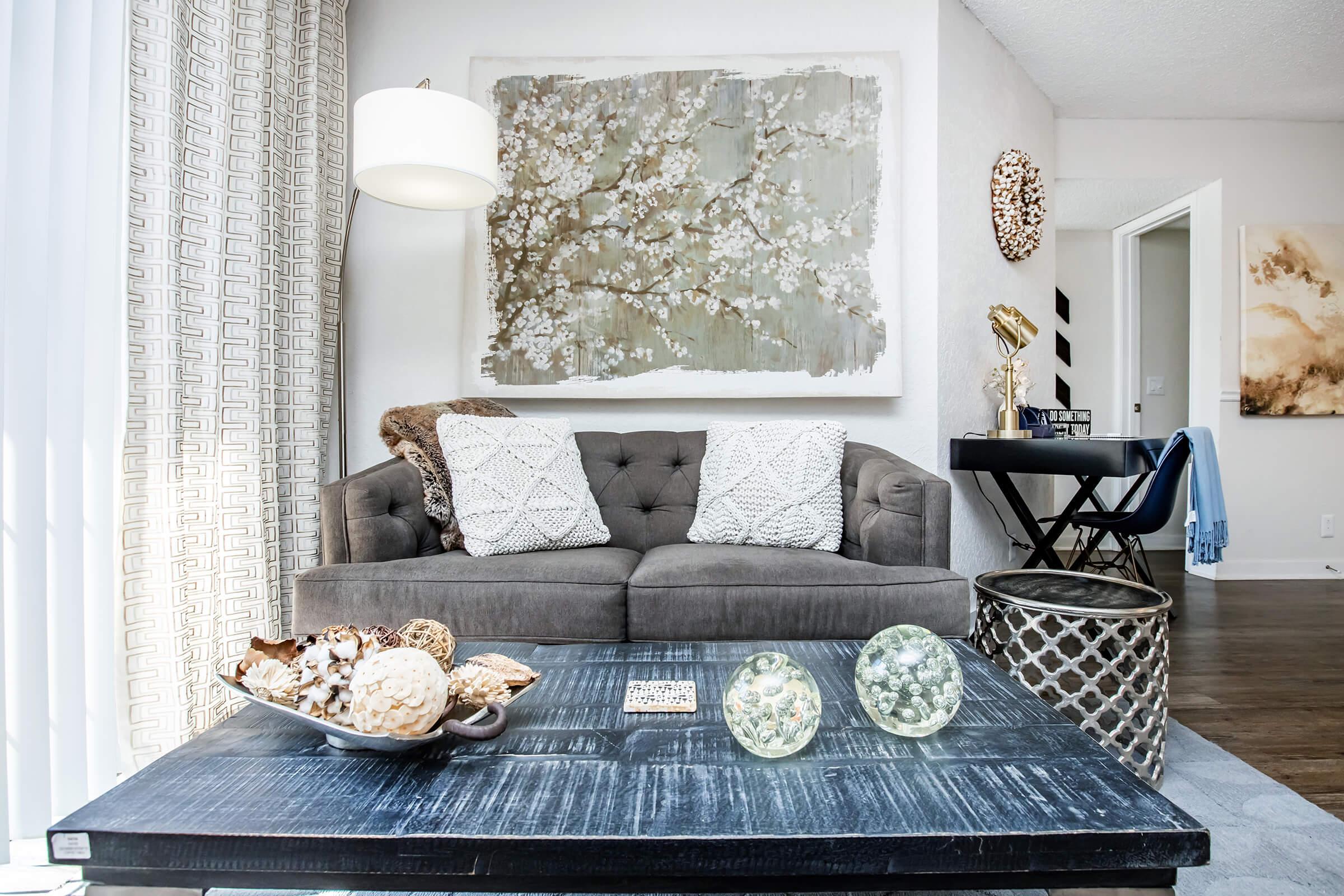 A cozy living room scene featuring a grey tufted sofa adorned with patterned pillows. In front of the sofa, there is a dark wooden coffee table with decorative items, including a bowl of natural elements and glass orbs. The space is brightened by a hanging lamp, with art on the walls and a small table visible in the background.