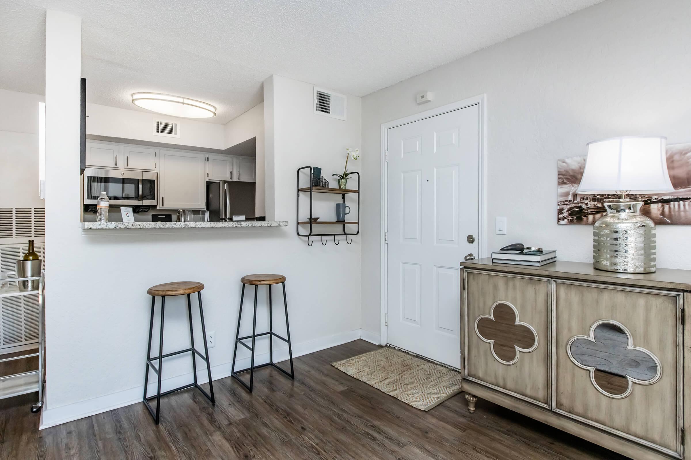 Interior view of a modern apartment entryway featuring a console table with decorative accents, two barstools at a kitchen island, and a front door. Natural light illuminates the space, highlighting the stylish decor, which includes a lamp and wall art. The overall aesthetic is contemporary and welcoming.