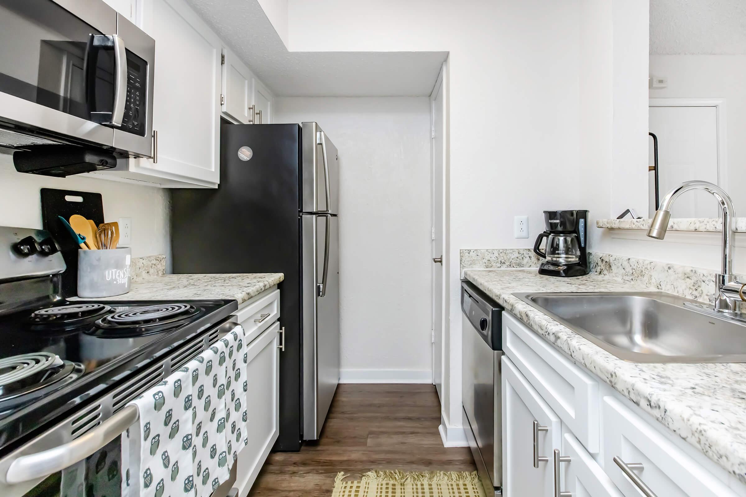Modern kitchen featuring white cabinets, granite countertops, and stainless steel appliances. There's a black refrigerator, a coffee maker, and an oven with a stovetop. Natural light illuminates the space, highlighting a patterned dish towel and a textured runner on the floor.
