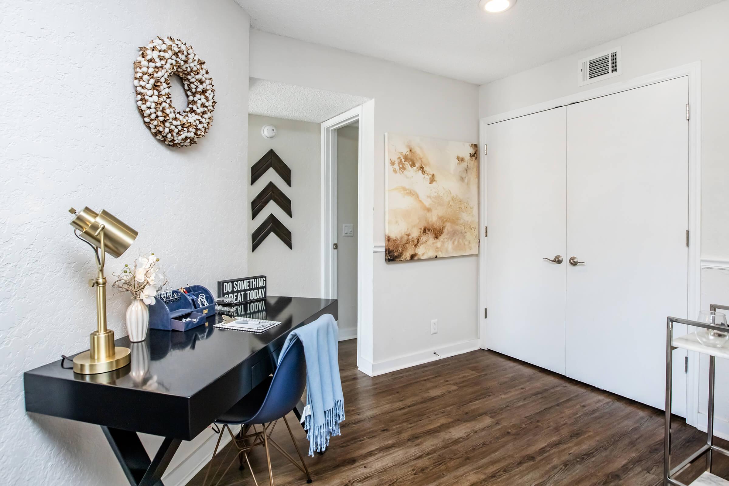 A modern interior office space featuring a black desk with a gold lamp, decorative items, and a blue throw. The wall is adorned with a wreath and an abstract art piece. A white door and wall accents complete the layout, with wooden flooring adding warmth to the room.