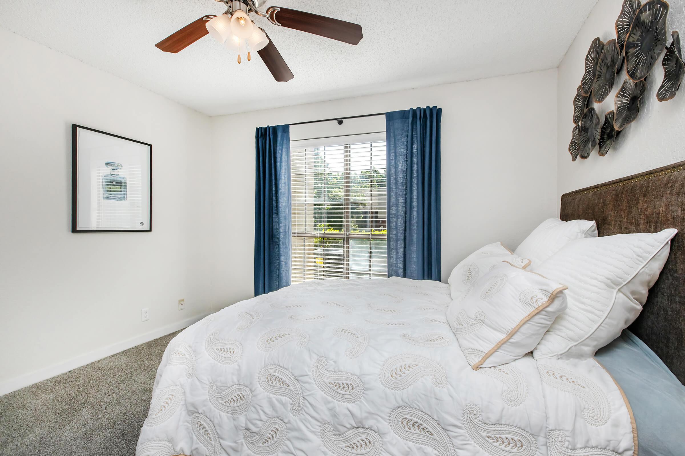 A cozy bedroom featuring a queen-sized bed with white bedding, a decorative wall art piece, and a ceiling fan. The room has large windows covered with blue curtains, allowing natural light to enter, and a carpeted floor. A framed picture hangs on the wall, adding to the room's inviting ambiance.