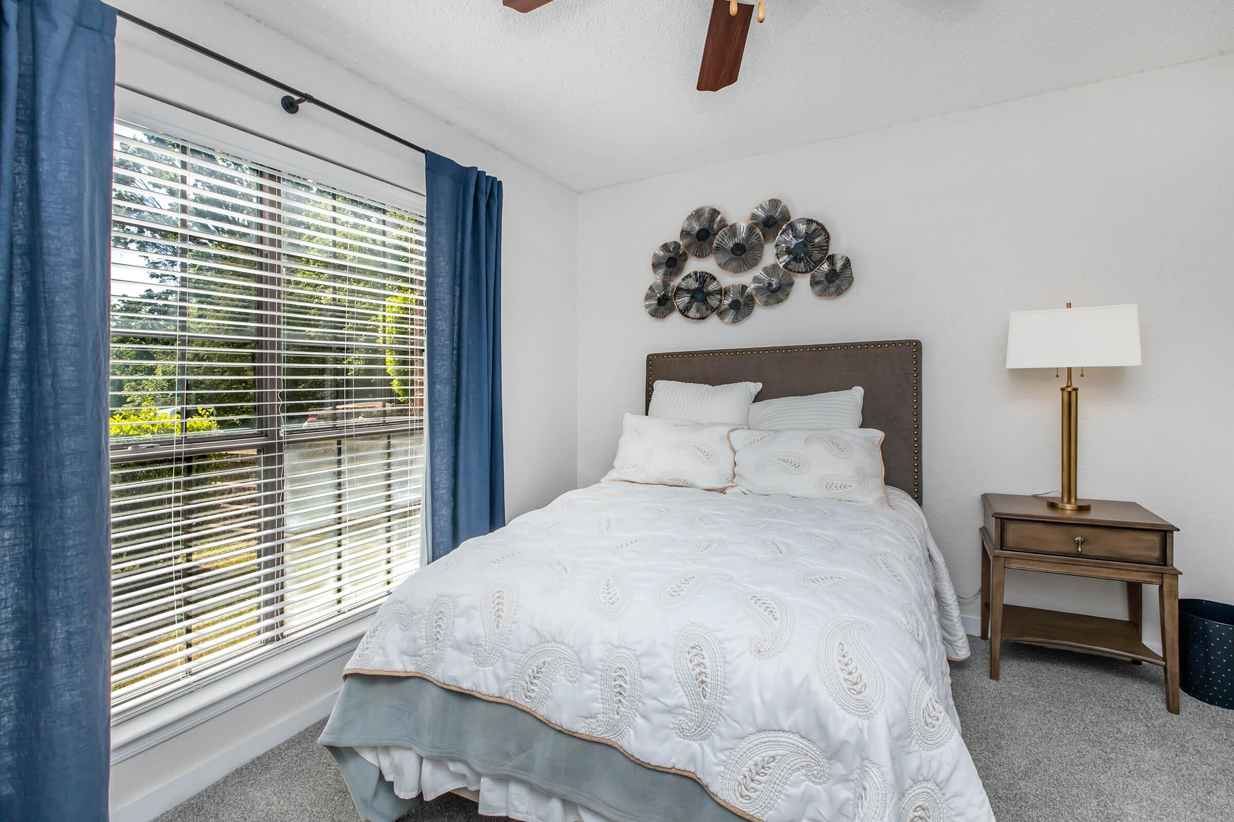 Cozy bedroom featuring a double bed with elegant bedding and decorative pillows. A bedside table with a lamp is beside the bed. Large window adorned with blue curtains offers natural light and a view of greenery outside. Soft carpet flooring enhances the inviting atmosphere of the space.