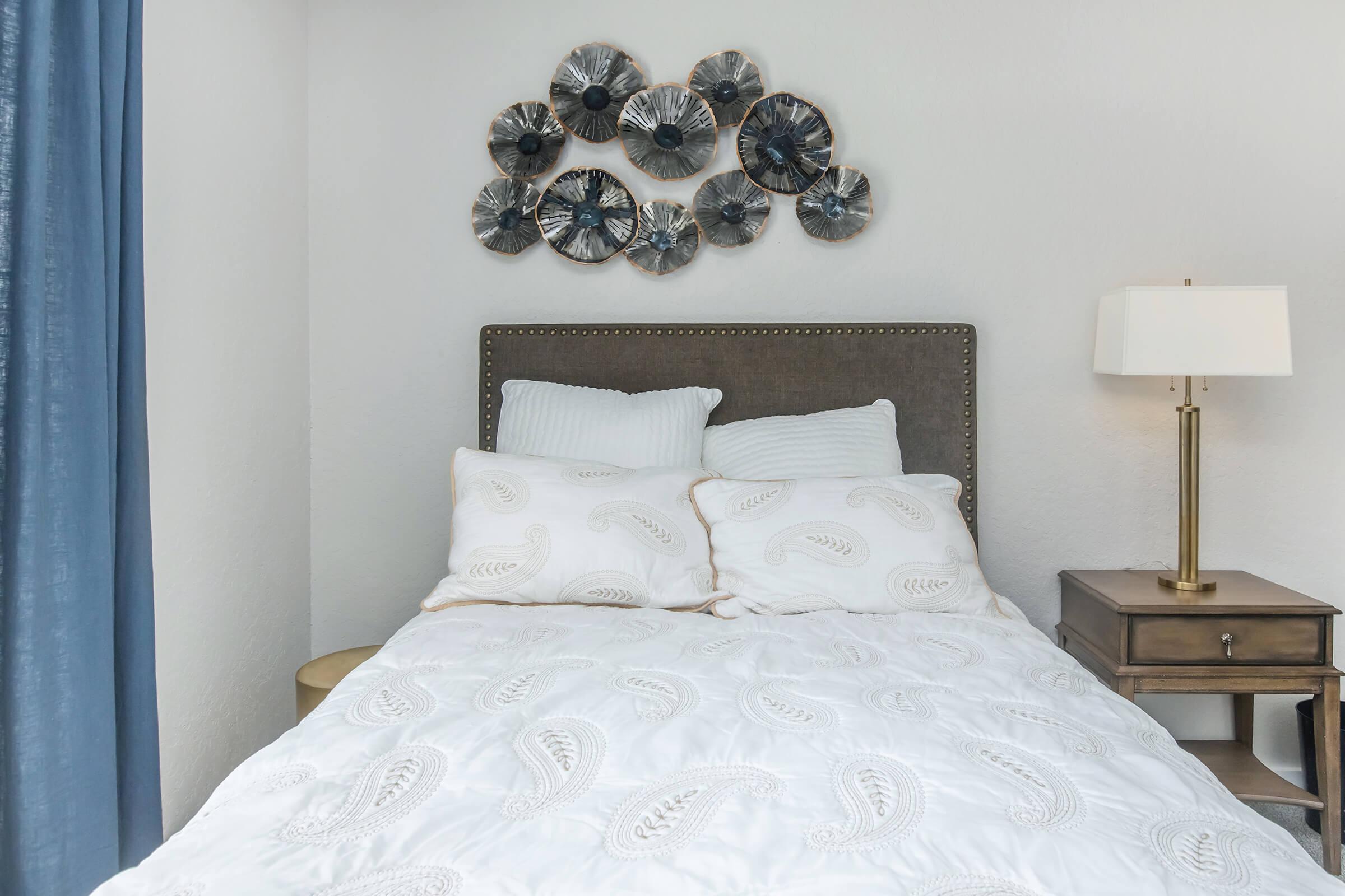 A cozy bedroom featuring a neatly made bed with white bedding and decorative pillows. Above the bed, a circular wall art display made of various textured elements is mounted. To the side, there's a nightstand with a lamp and decorative items, complemented by a blue curtain in the background.