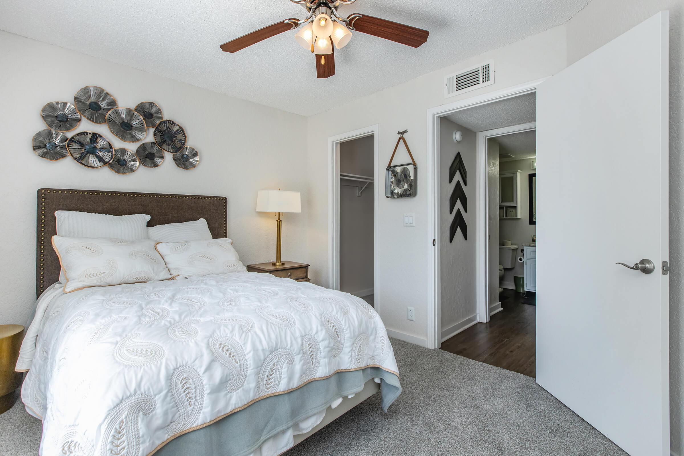 A cozy bedroom featuring a queen-sized bed with a decorative white and cream comforter. There’s a bedside lamp and a wall adorned with circular metal art. The room has a ceiling fan and a door leading to a bathroom, with light-colored walls and carpeted flooring for a warm and inviting atmosphere.