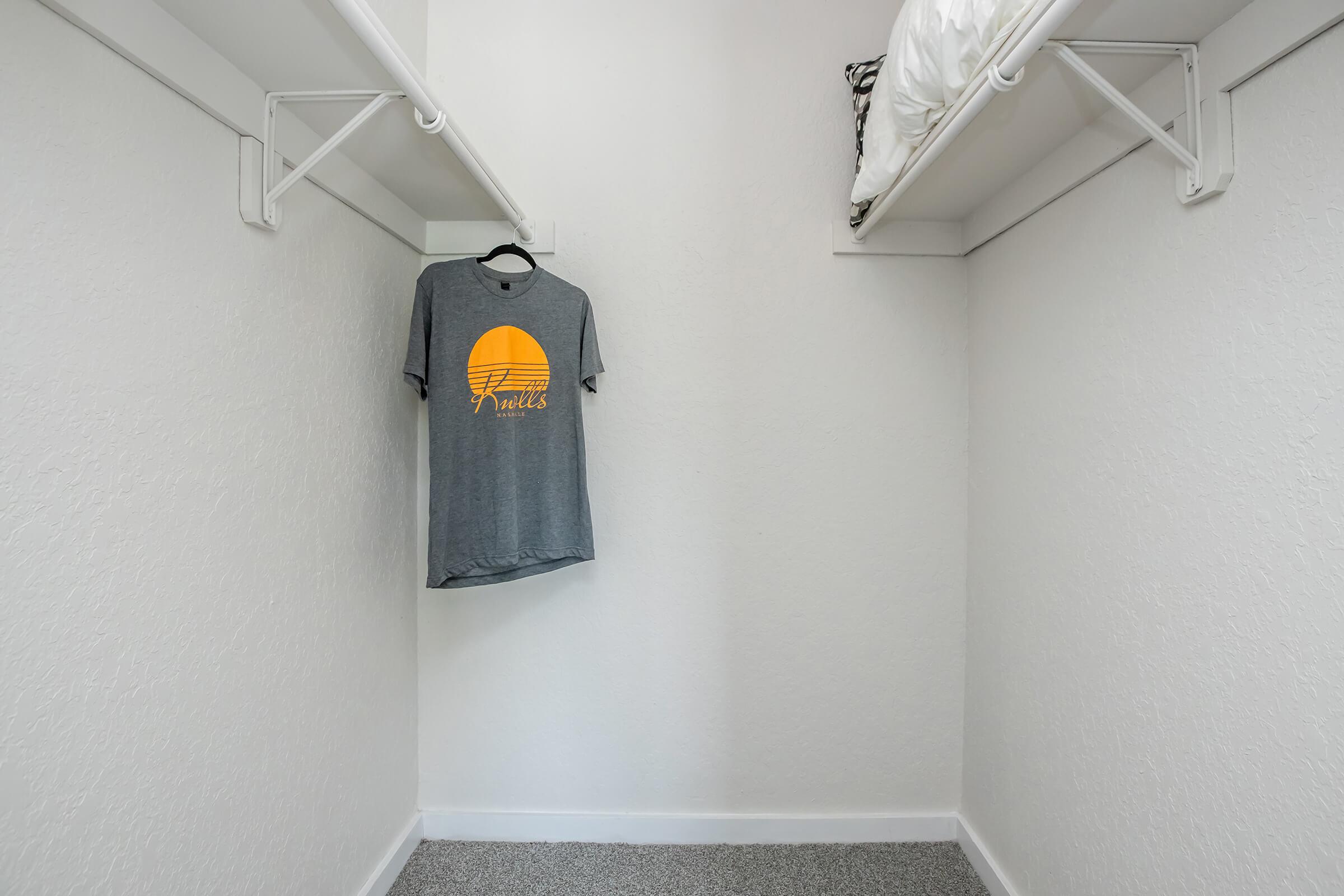 A spacious, minimally decorated closet with white walls and gray carpet. A gray t-shirt with a yellow sun graphic hangs on a metal hanger from a shelf, while the opposite shelf holds a neatly folded comforter.