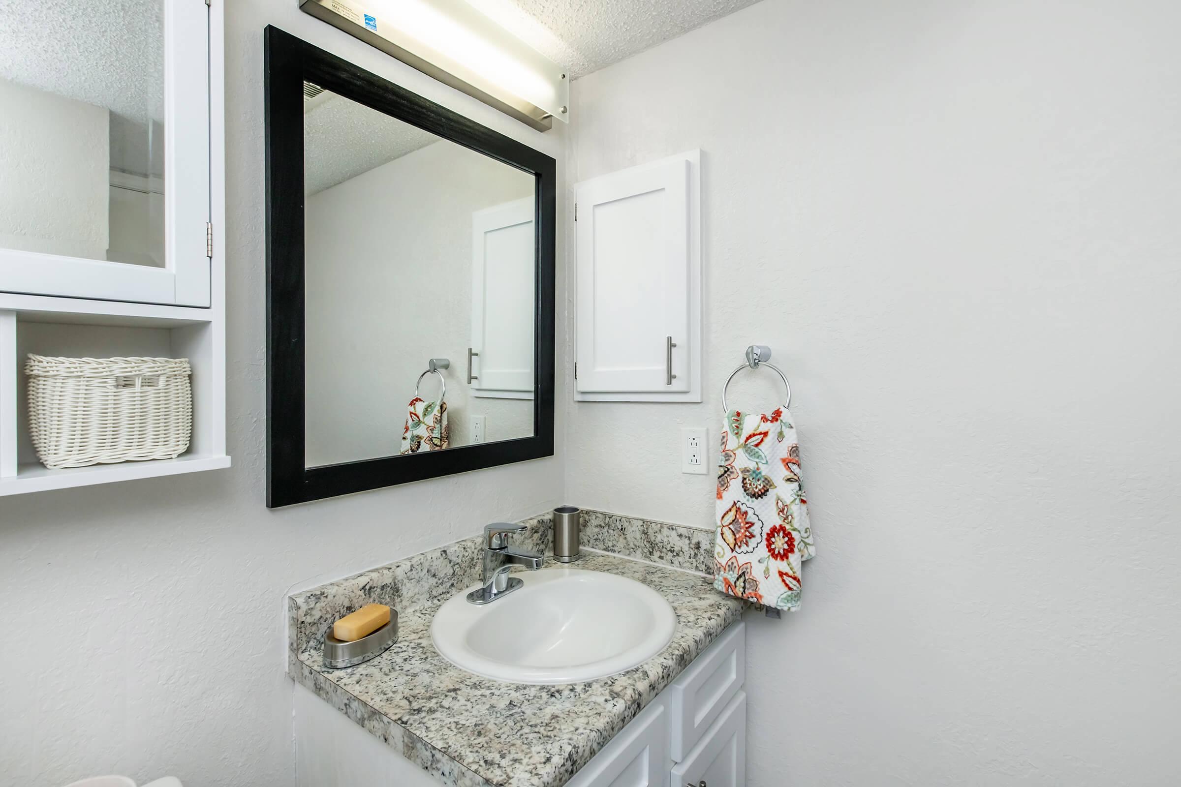 A modern bathroom featuring a granite countertop with a circular sink, a wall-mounted soap dispenser, and a decorative hand towel hanging on a silver holder. There's a large black framed mirror above the sink, with a bright light fixture above it. White cabinetry and a woven basket add storage and style to the space.