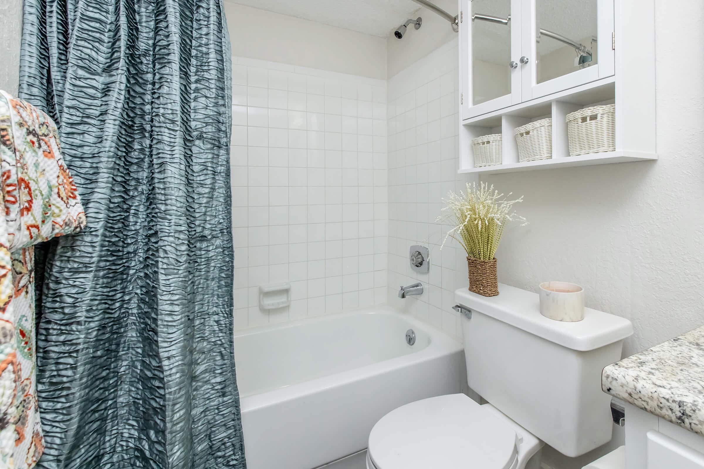 A clean and tidy bathroom featuring a white tub with a shower curtain, a toilet, and a sink with a granite countertop. There are shelves above the toilet with decorative baskets and a small vase of faux flowers. The walls are light-colored, enhancing the spacious feel of the room.