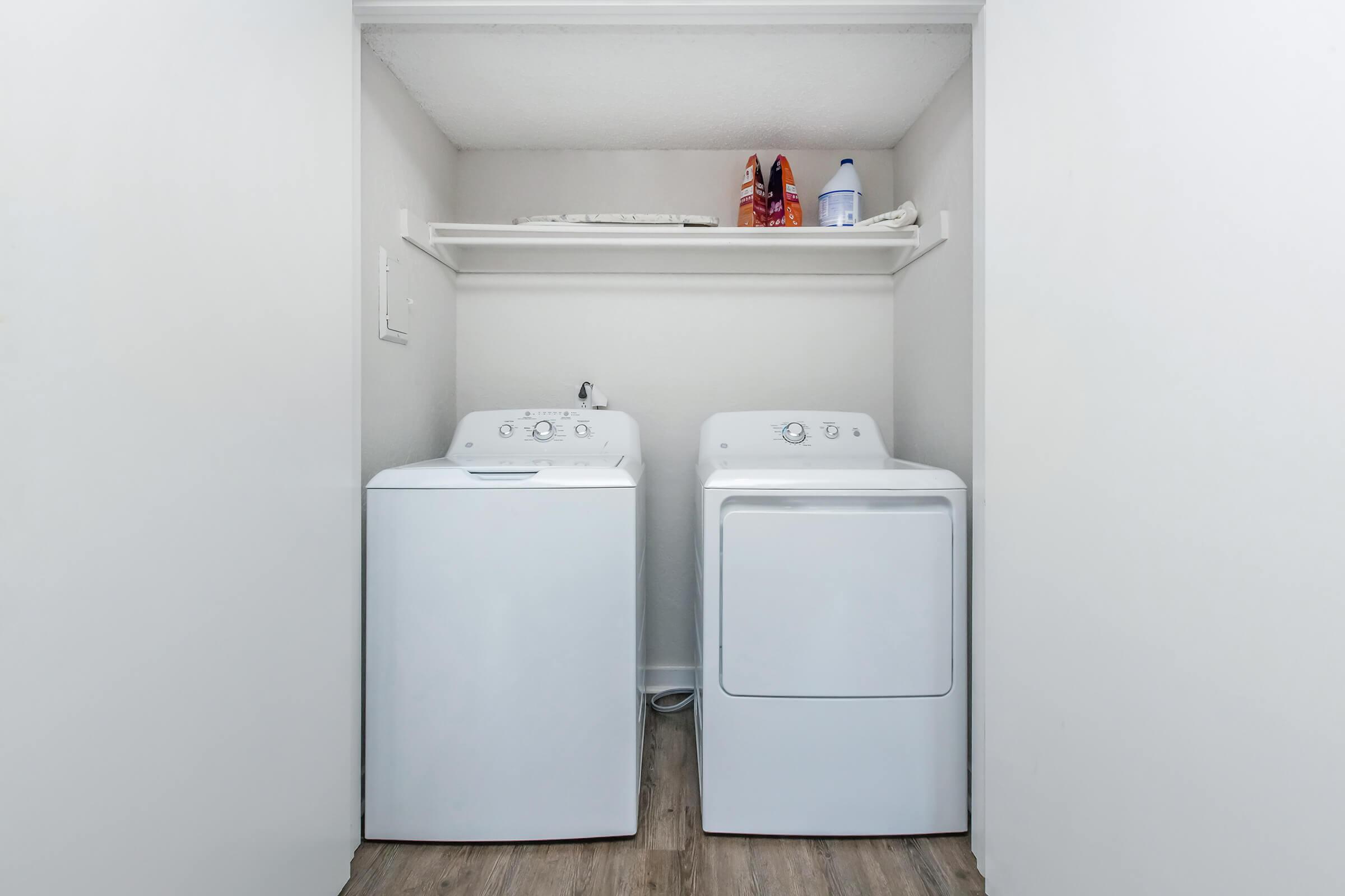 A clean laundry room featuring a white washing machine and a white dryer side by side. Above them, a shelf holds a bottle of detergent and a pair of red shoes. The walls are light-colored, and the flooring is a wooden laminate. The space is well-organized and tidy.