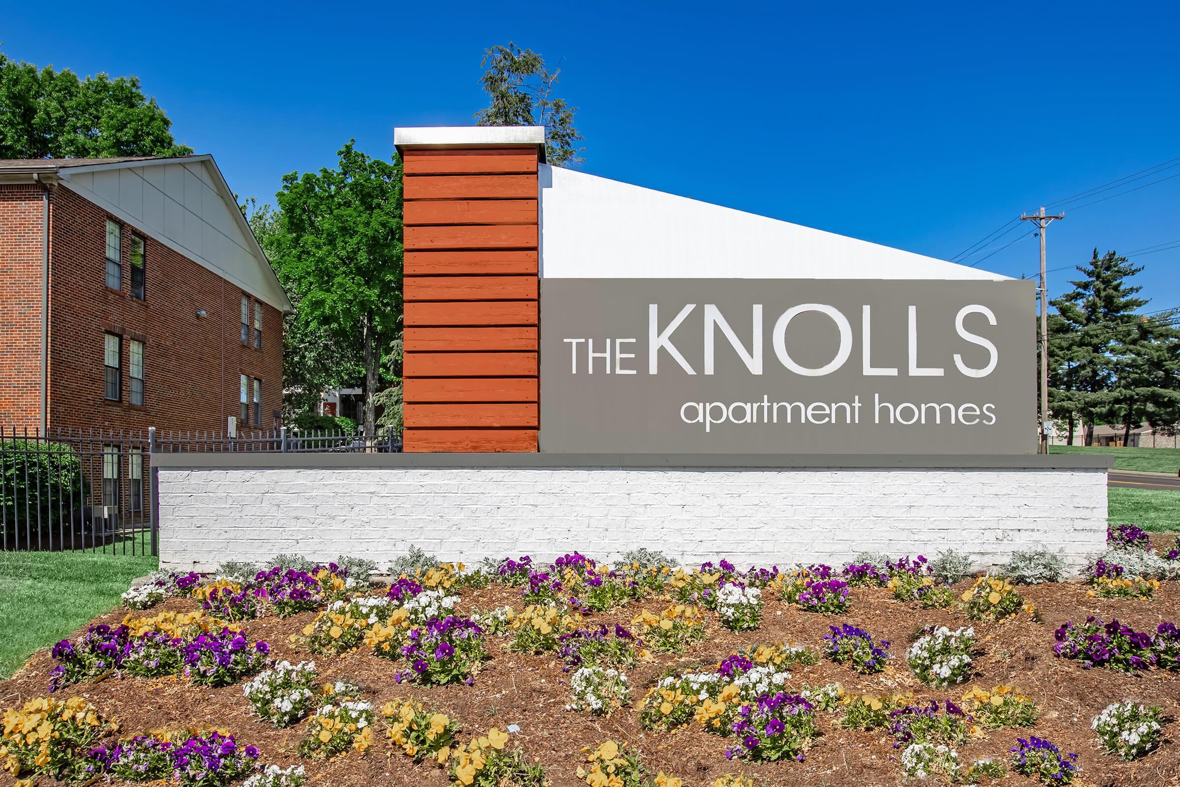 Sign for The Knolls Apartment Homes featuring a modern design with a red and gray color scheme. The sign is placed in front of a landscaped area with colorful flowers and greenery under a clear blue sky.