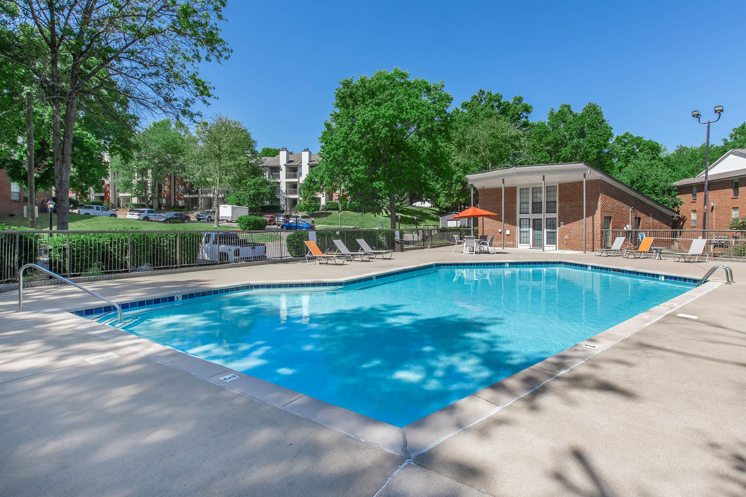 A clear blue swimming pool surrounded by a concrete deck, with lounge chairs and orange umbrellas. Lush green trees and apartment buildings are visible in the background under a bright blue sky.