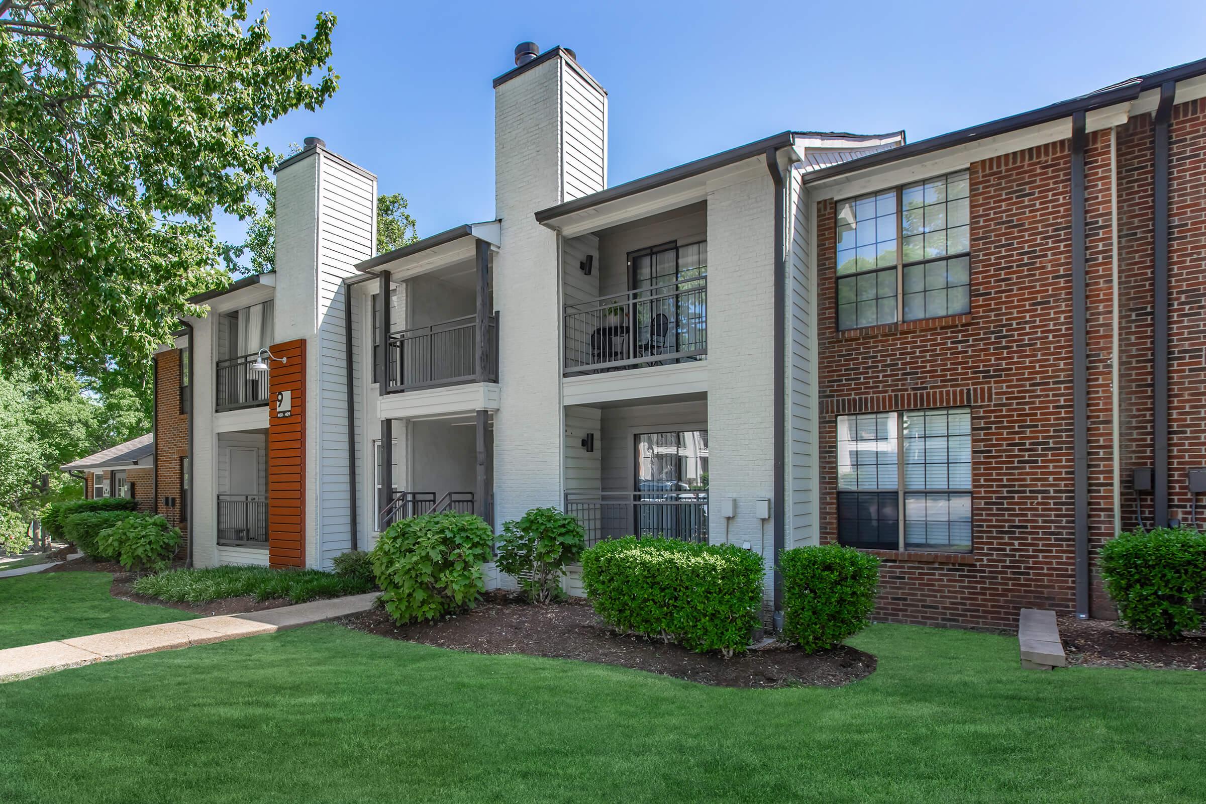 A modern apartment building featuring a mix of white and brick facades, surrounded by lush greenery. The scene includes well-maintained lawns and balconies, with clear blue skies in the background, suggesting a pleasant residential environment.