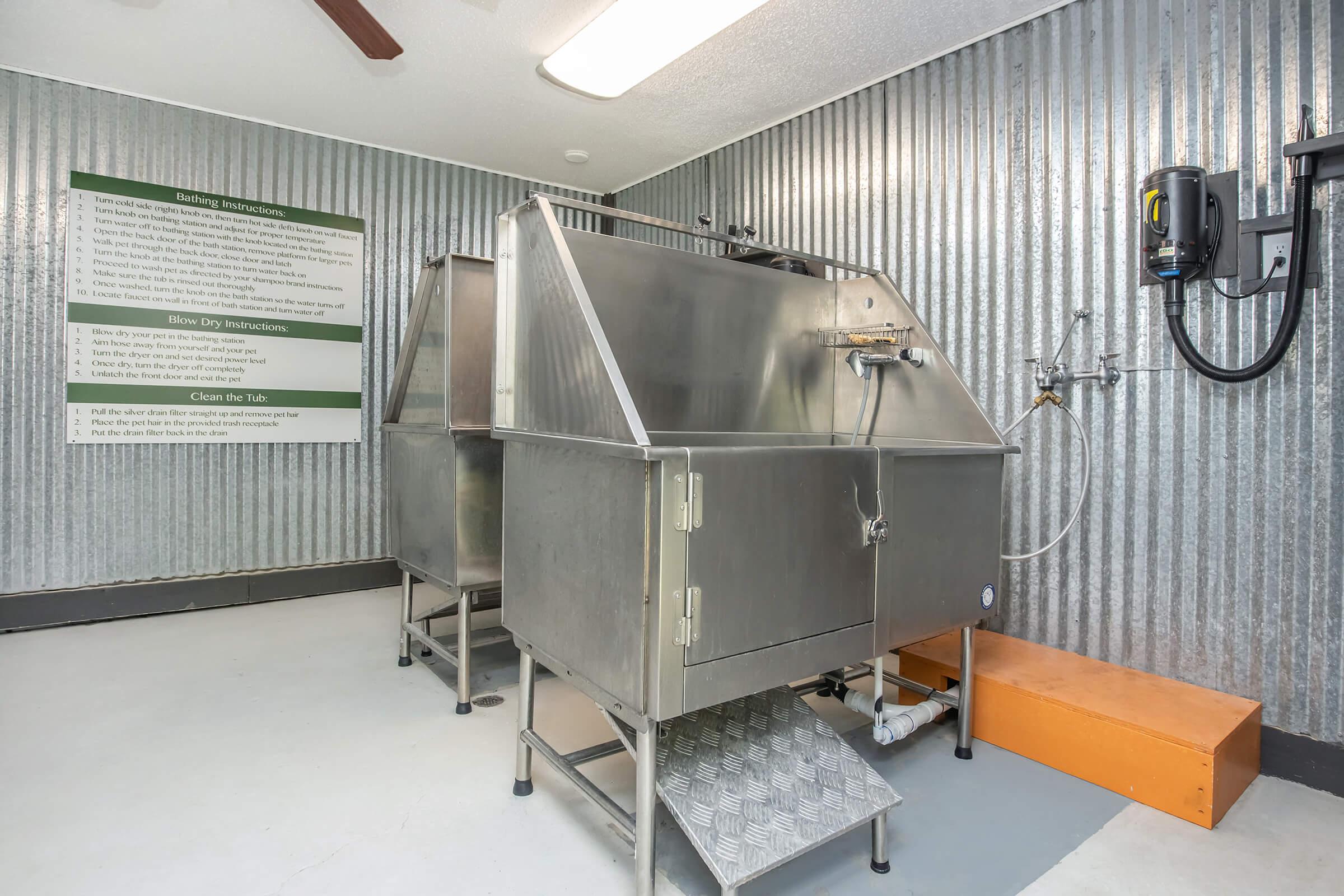 Two stainless steel dog bathing tubs positioned in a room with corrugated metal walls. A sign with instructions is displayed on the wall behind the tubs. The floor is light-colored, and there’s a mounted drying device on the wall. The space is designed for pet grooming.