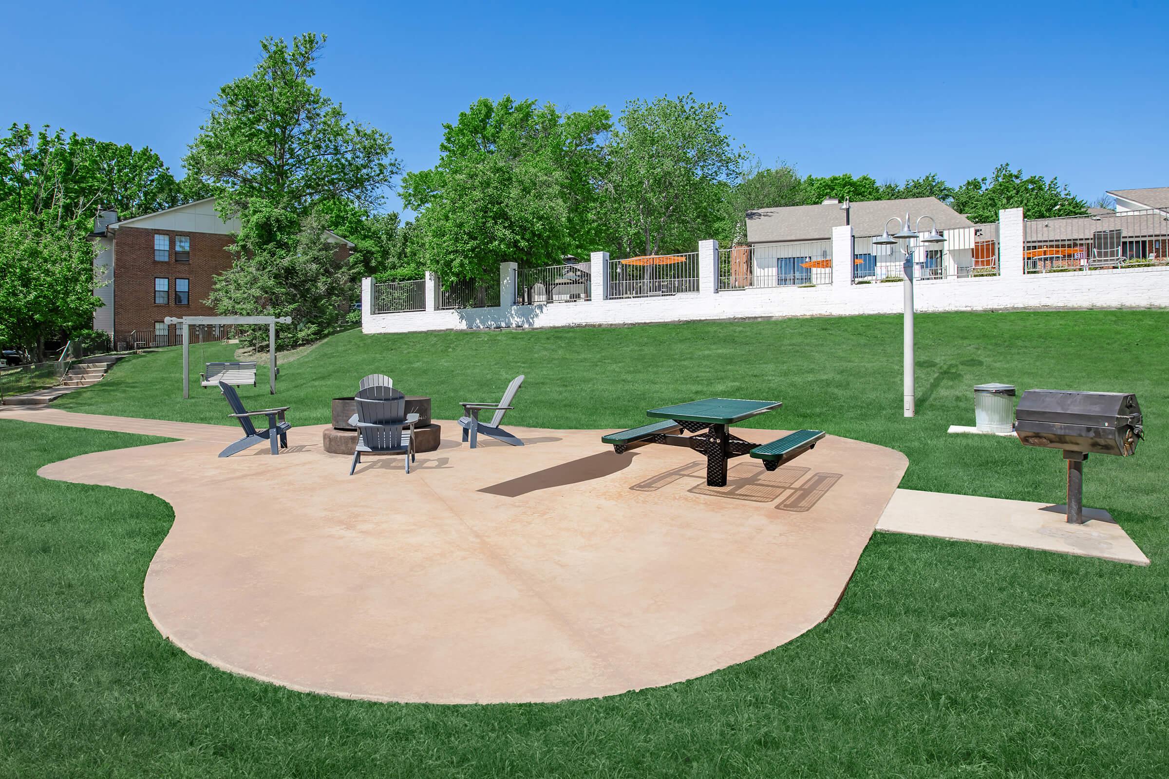 A grassy outdoor area featuring a circular concrete patio with picnic tables, adirondack chairs, a fire pit, and a grill. In the background, there are trees and buildings, creating a welcoming space for gatherings and recreation. Clear blue sky overhead enhances the inviting atmosphere.