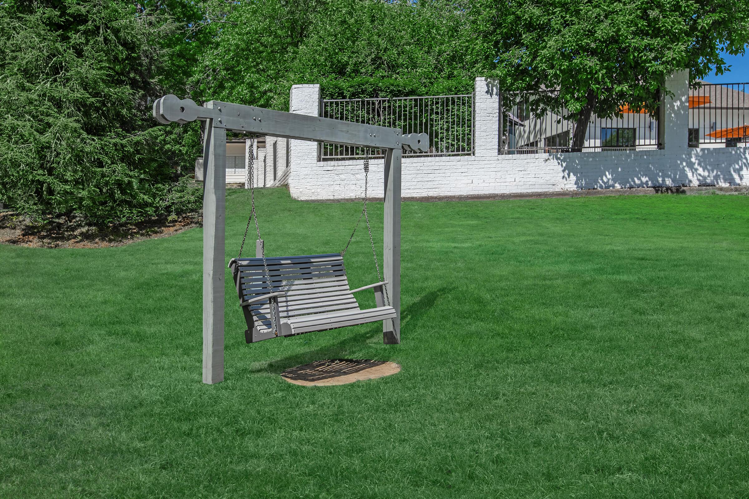 A wooden swing set on a grassy lawn, surrounded by trees and a white brick wall in the background. The swing is attached with chains and positioned on a circular wooden platform. The setting is bright and sunny, creating a peaceful outdoor atmosphere.