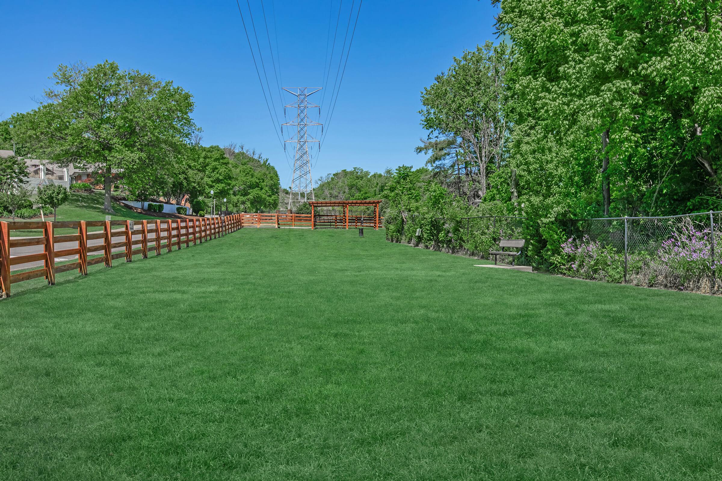 A spacious, green park area featuring lush grass, a wooden fence lining the path, and a shaded gazebo. There are tall trees along the perimeter, and power lines stretch across the clear blue sky. A bench is visible near the fence, providing a place to sit and enjoy the scenery.