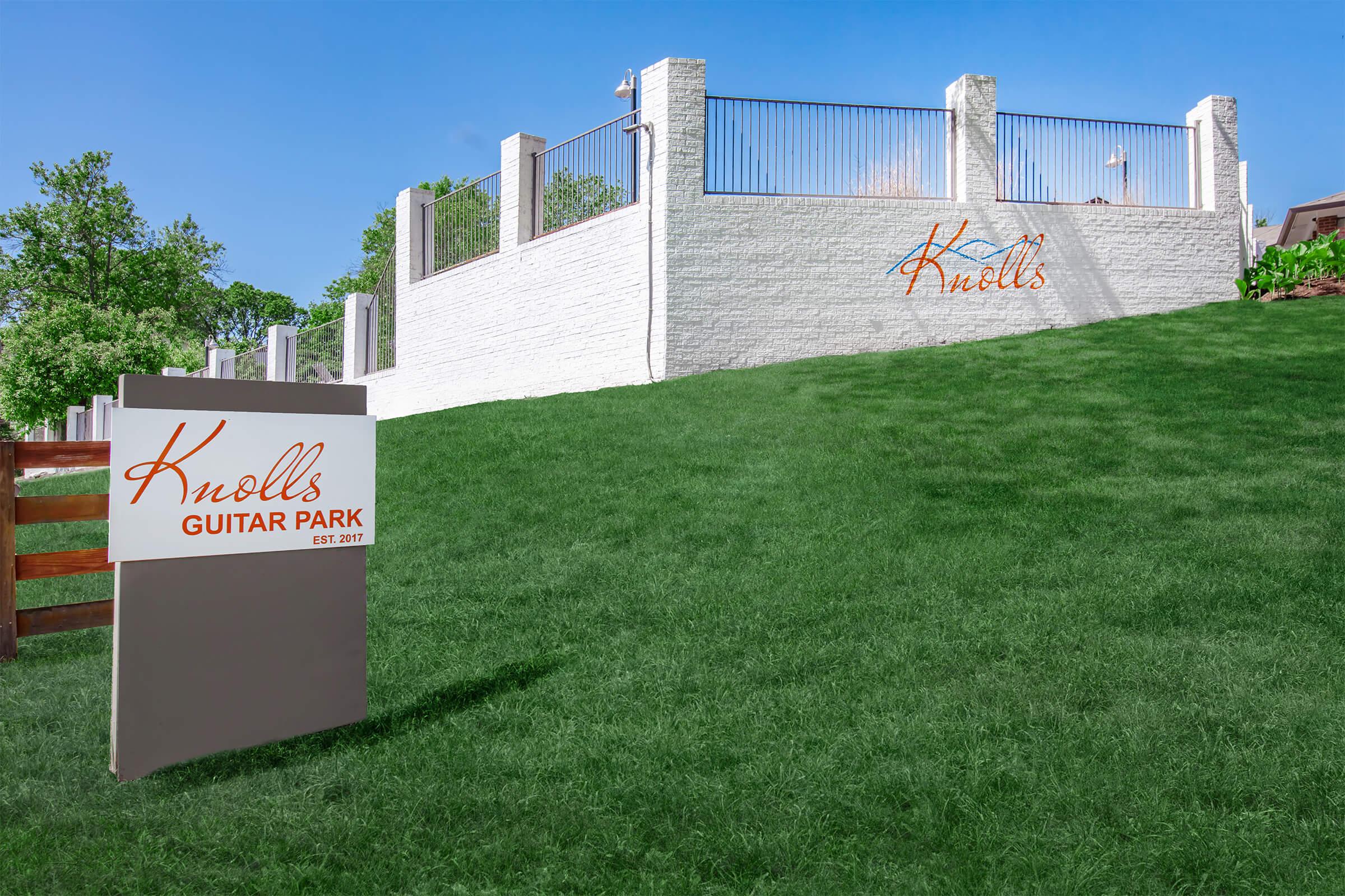 Sign for Knolls Guitar Park in front of a green grassy area with a white wall and a fence in the background. The sign features the text "Knolls Guitar Park" and "EST. 2017." The setting appears to be bright and welcoming, suggesting a park dedicated to guitar enthusiasts.