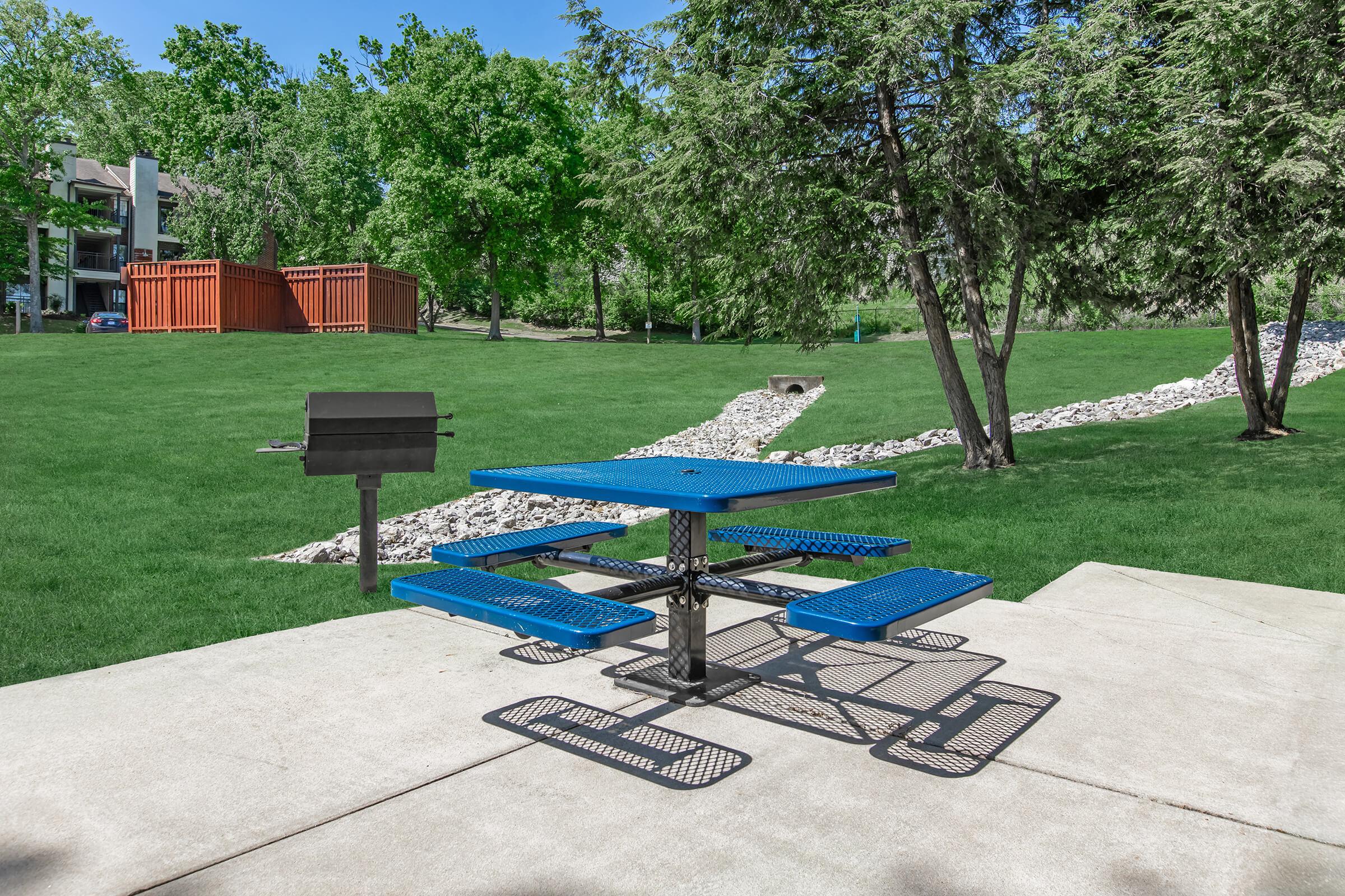 A blue picnic table with attached benches sits on a concrete slab in a spacious green park. Nearby, there is a charcoal grill and a landscaped area featuring a stone path winding through grass and trees. Sunlight brightens the scene, creating a welcoming outdoor atmosphere.