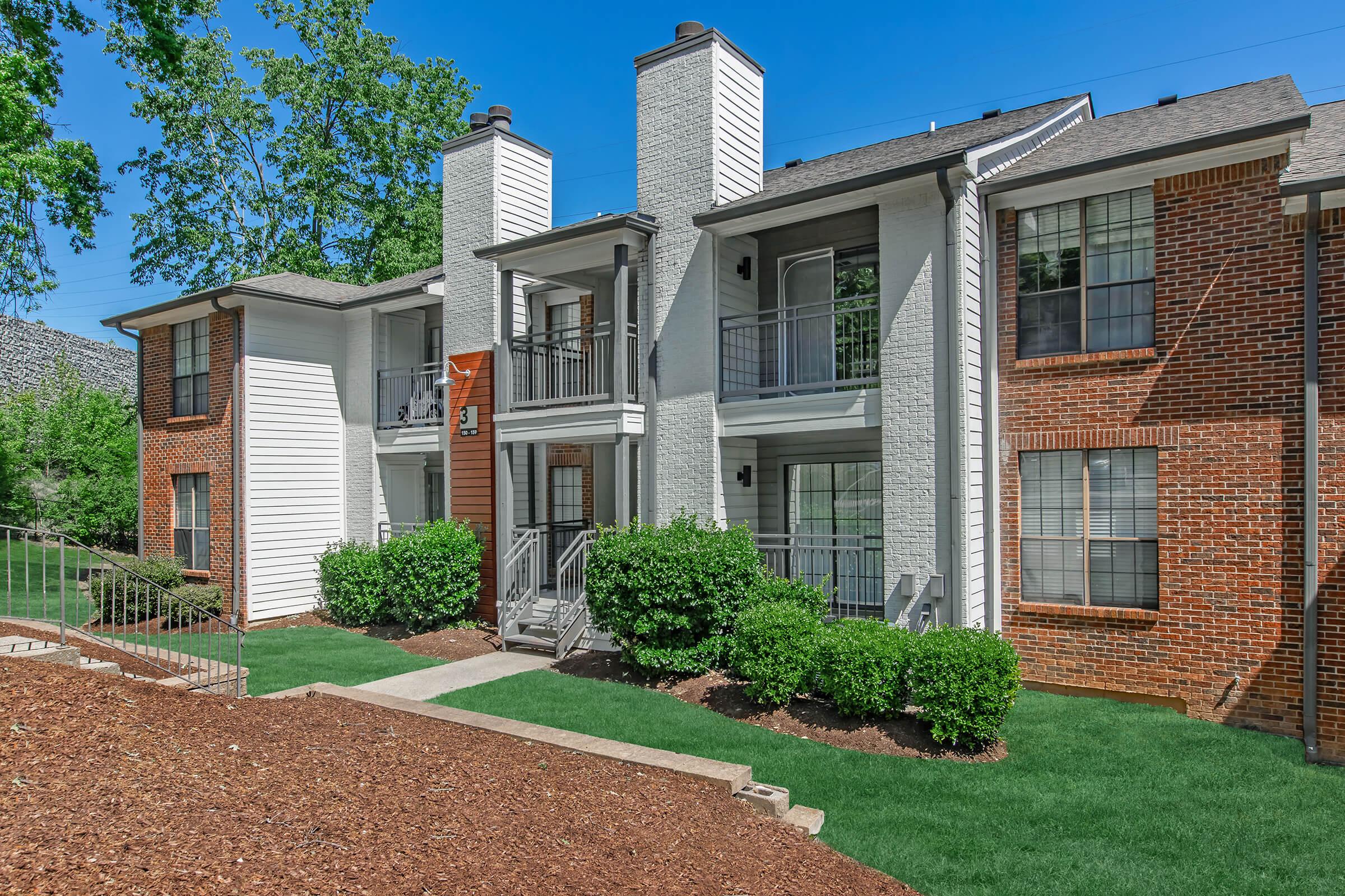 A view of a modern apartment complex featuring two building styles: one with brick siding and the other with white siding. The buildings have multiple balconies and large windows, surrounded by well-manicured lawns and small shrubs. The sky is clear and blue, indicating a sunny day.