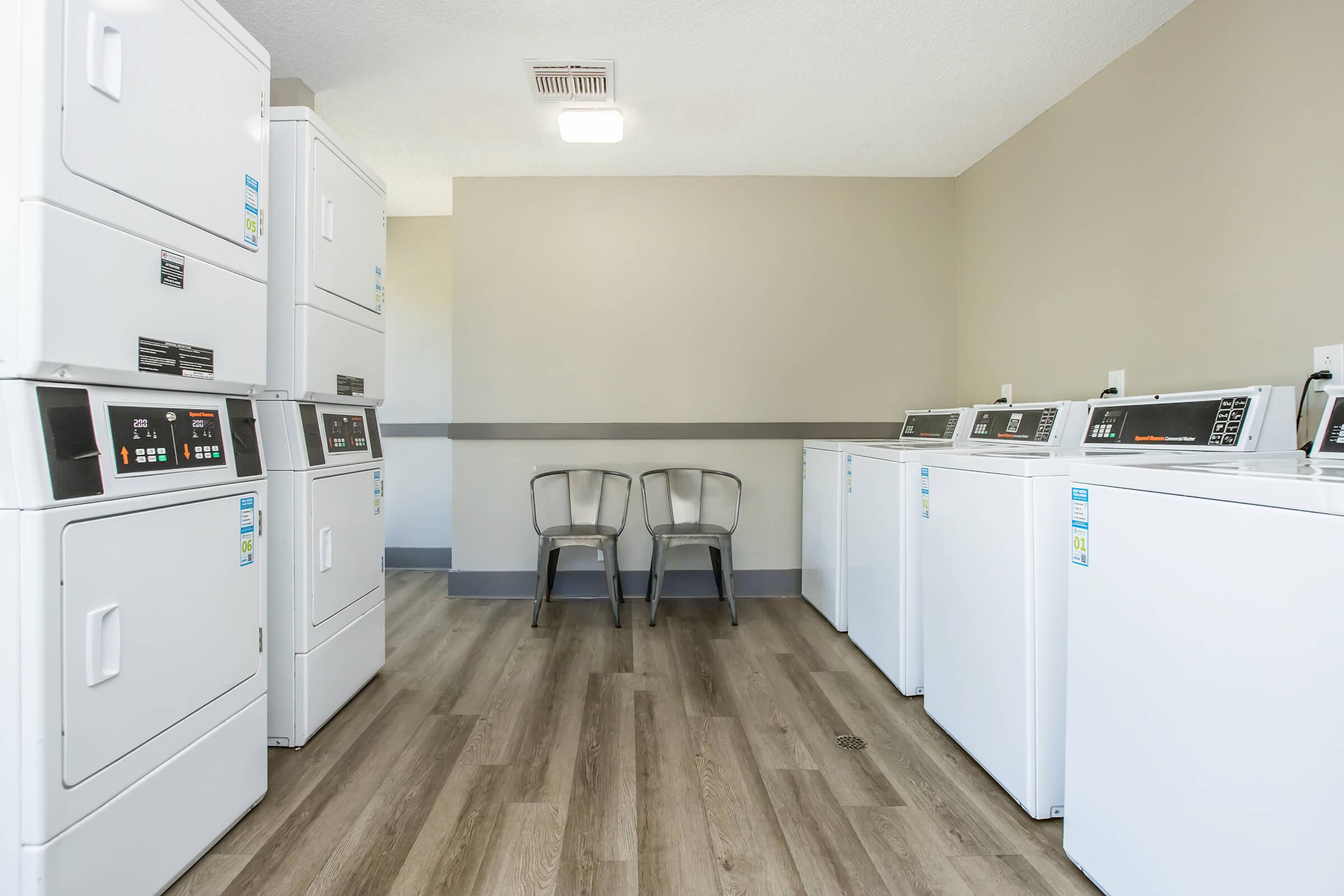 A clean and modern laundry room featuring several white washing machines and dryers, arranged neatly. There are two metal chairs against the wall, and the room has light-colored walls and wooden flooring, creating a bright and inviting atmosphere.