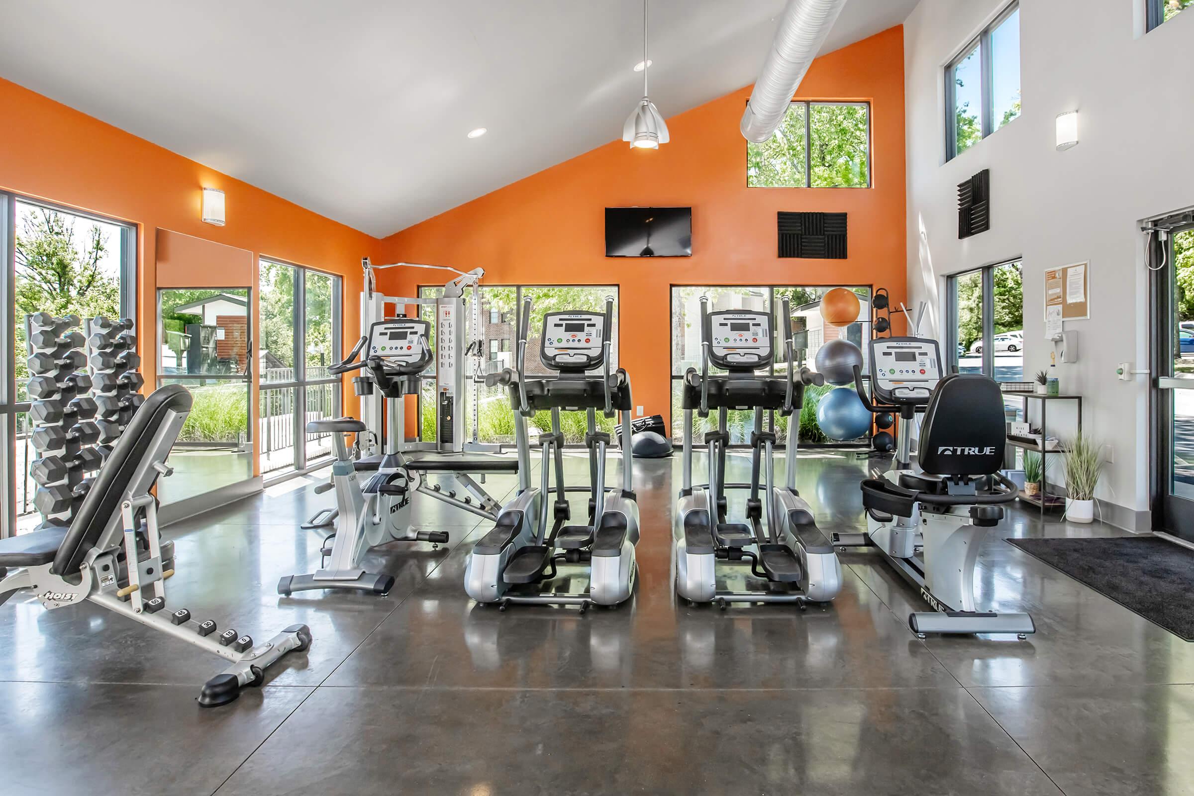 A modern gym interior featuring several exercise machines, including treadmills and stationary bikes. Large windows allow natural light in, and orange accent walls enhance the vibrant atmosphere. Exercise equipment, such as free weights and stability balls, is visible, promoting a lively fitness environment.