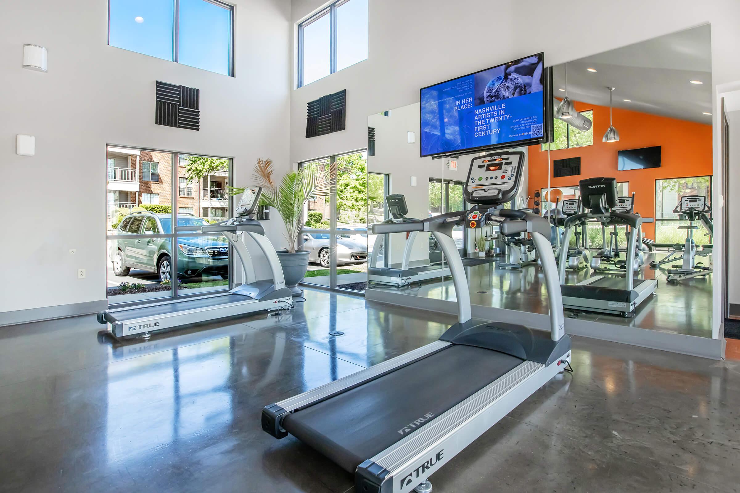 A modern fitness center featuring two treadmills with a large wall-mounted TV displaying fitness information. Bright windows let in natural light, and there are plants adding a touch of greenery. The space has sleek, polished floors and an inviting atmosphere, with a view of cars parked outside.