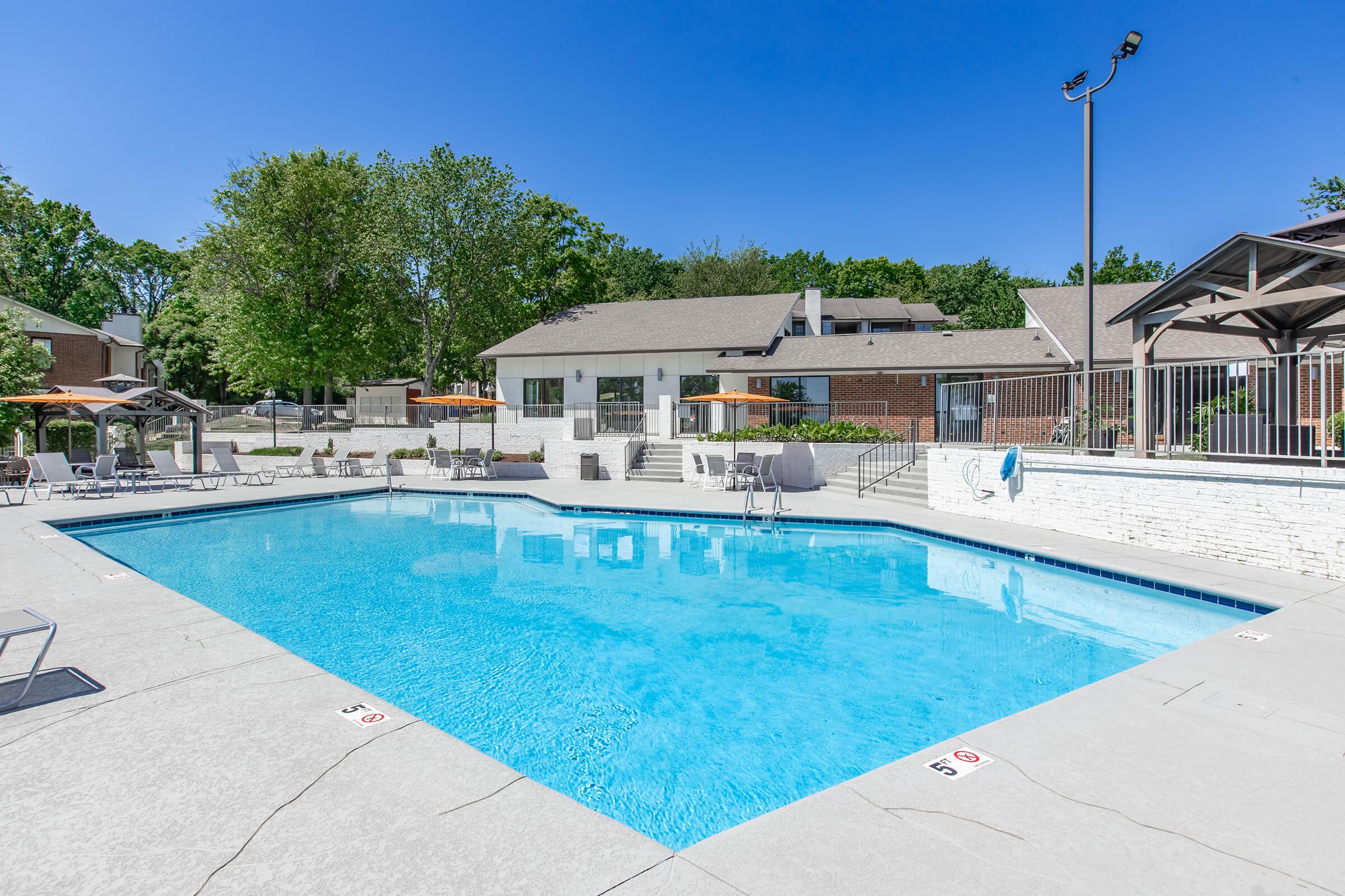 A bright, inviting outdoor swimming pool surrounded by lounge chairs and umbrellas. The pool area features a spacious deck and lush green trees and buildings in the background under a clear blue sky, perfect for relaxation and recreation.