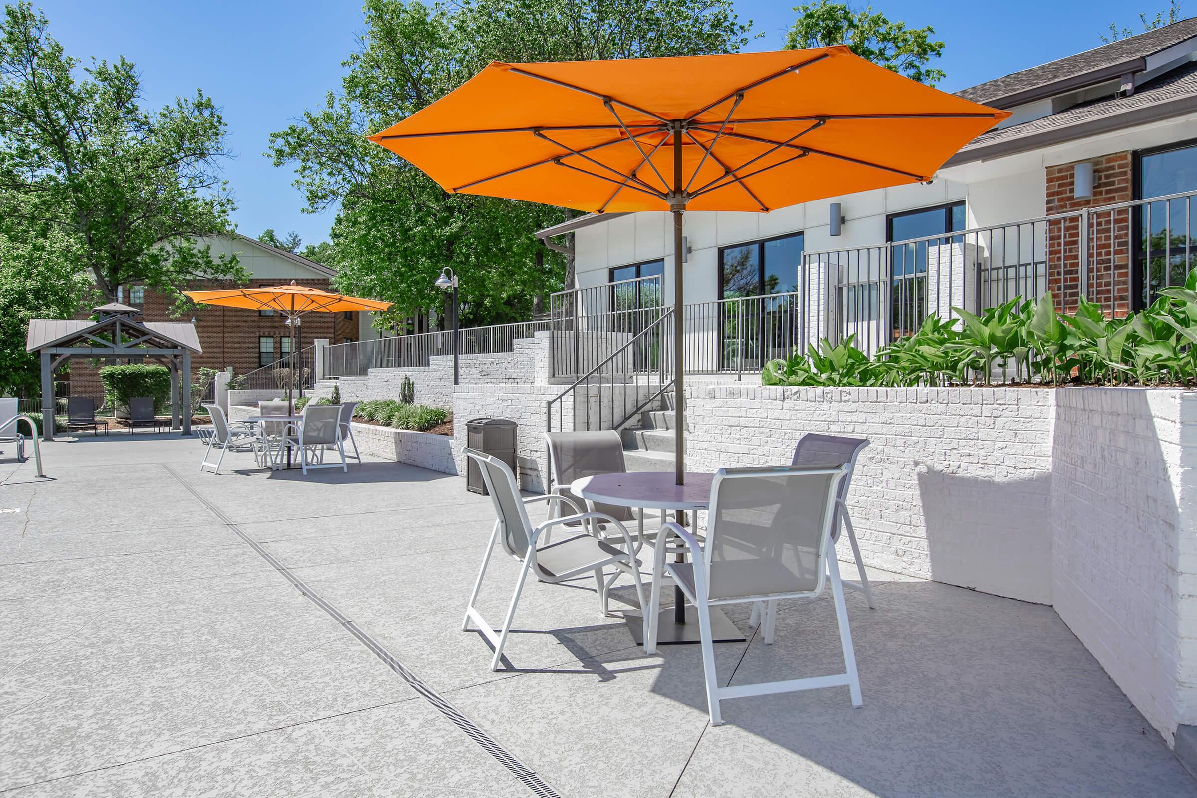 A sunny outdoor patio area featuring white brick walls, orange umbrellas over dining tables, and lush greenery. The space is well-lit and inviting, showcasing a modern design with comfortable seating arrangements, perfect for enjoying meals or relaxing outdoors.