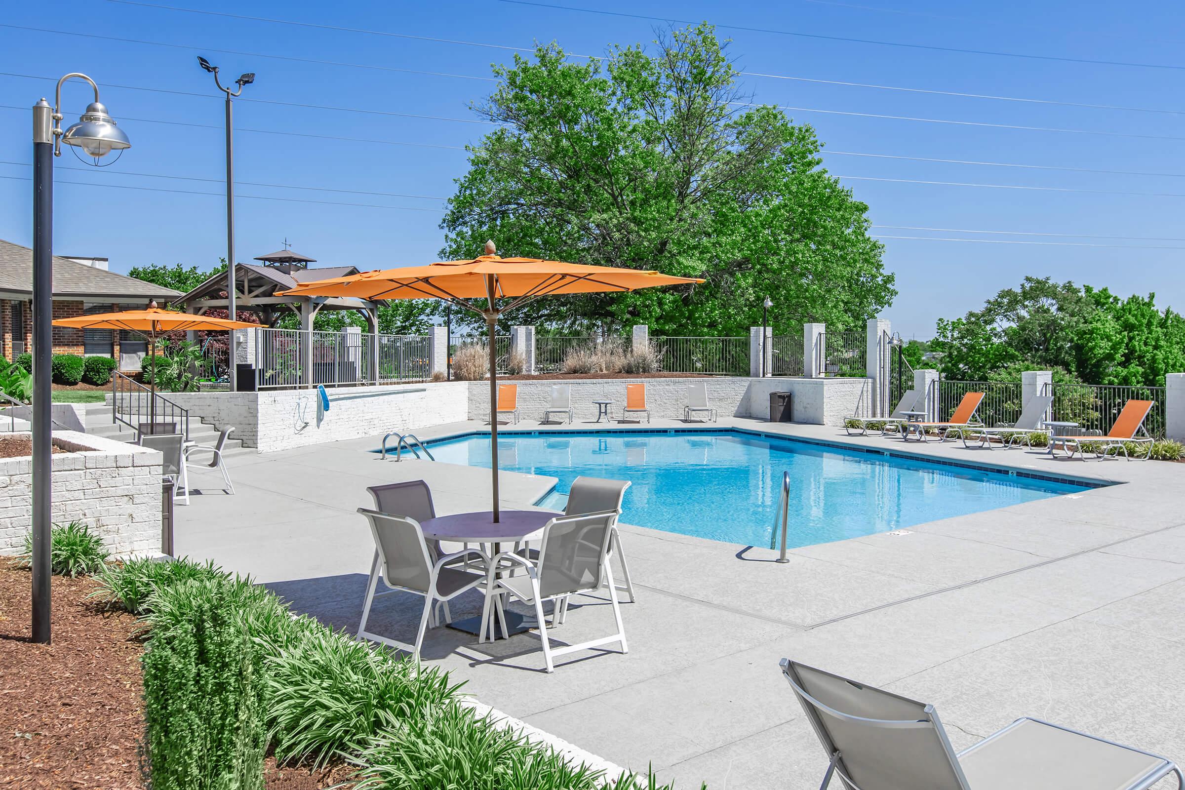A clear blue swimming pool surrounded by lounge chairs and orange umbrellas. Lush greenery and a landscaped area are visible in the background, with a bright blue sky overhead. The setting is inviting and ideal for relaxation.