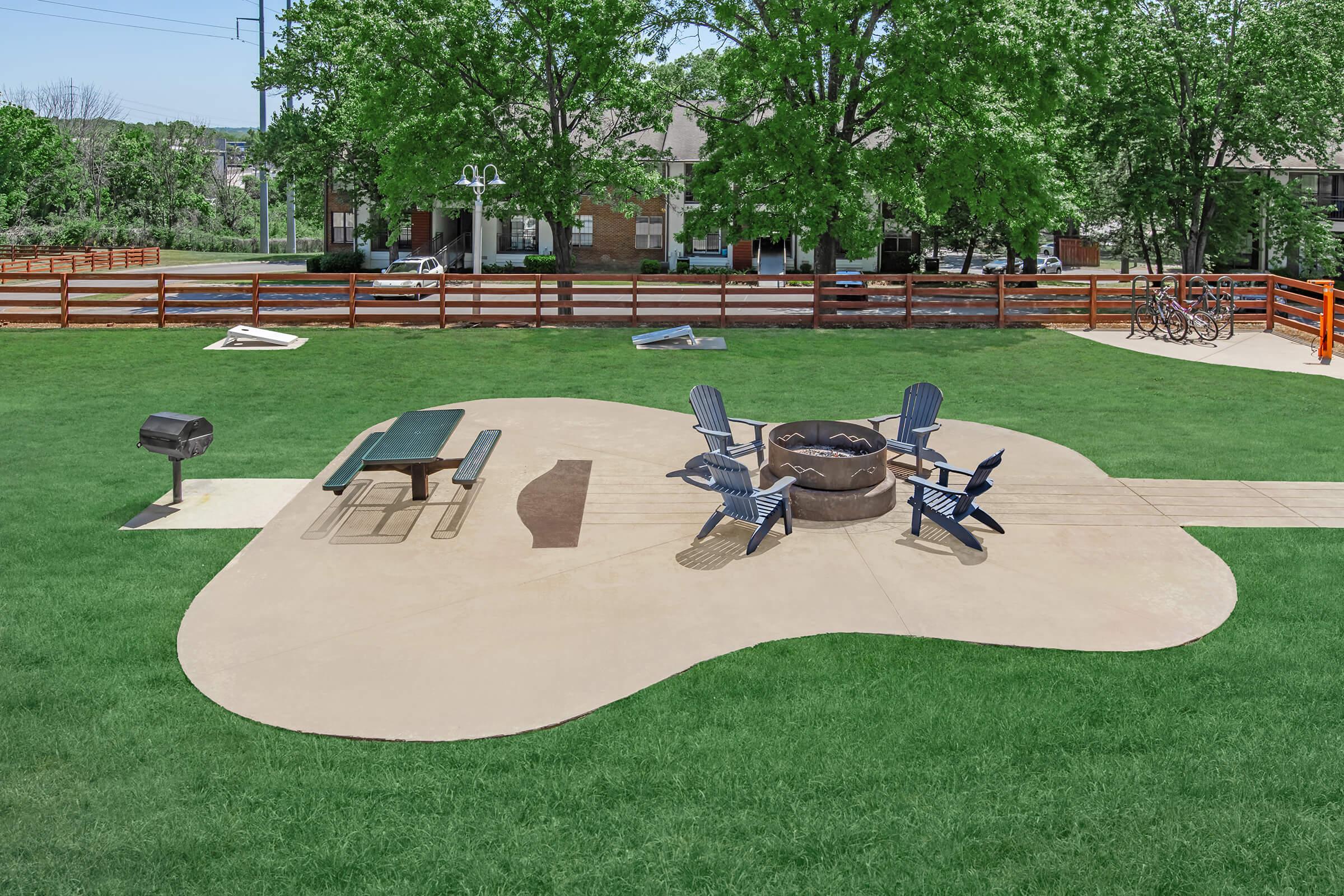 A well-maintained outdoor space featuring a unique guitar-shaped patio. It includes a picnic table, several blue chairs surrounding a fire pit, and a charcoal grill. Lush green grass surrounds the area, with trees in the background and a wooden fence enclosing the space.