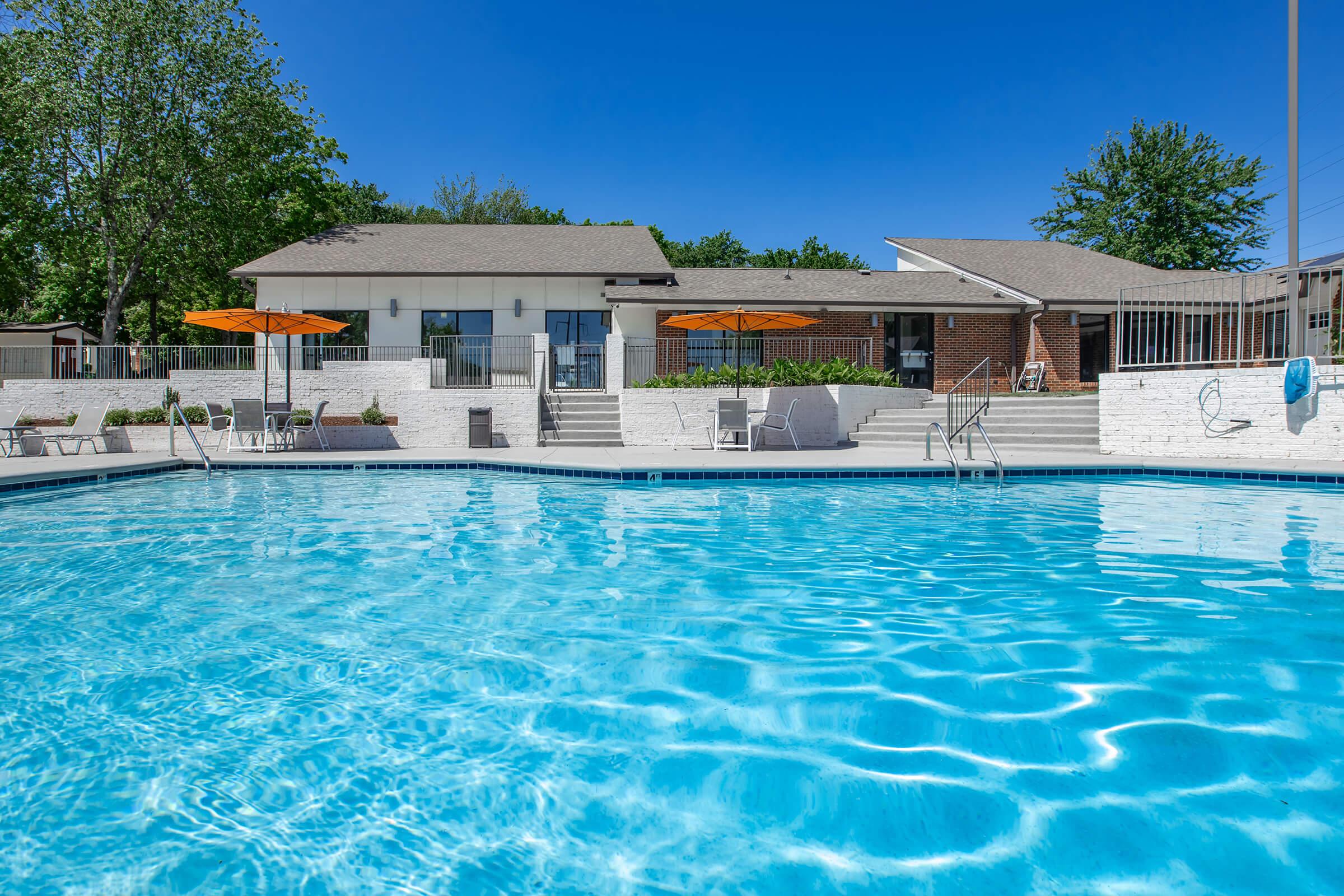 A clear blue swimming pool with a textured surface, surrounded by lounge chairs and orange umbrellas. In the background, a modern building with large windows and a patio area is visible, along with greenery from nearby trees under a bright blue sky.