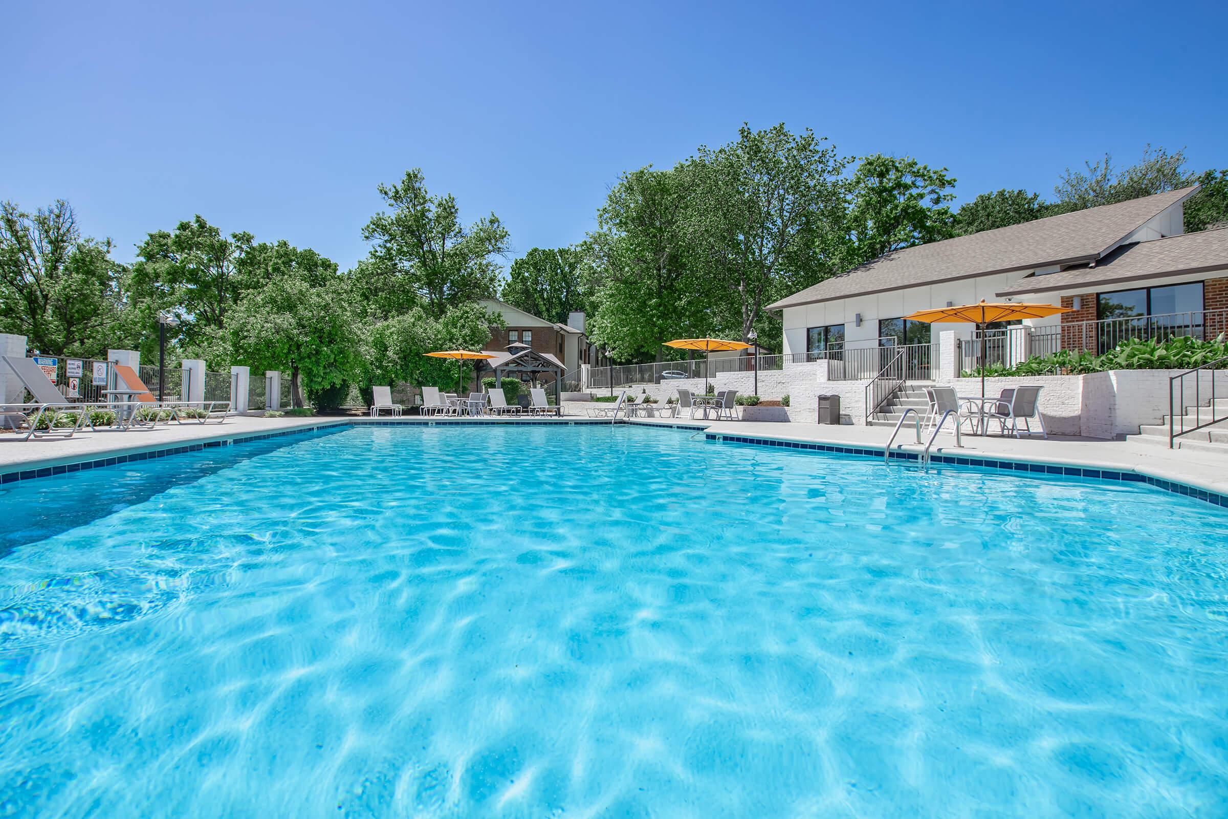 A bright, inviting swimming pool surrounded by lounge chairs and shaded by orange umbrellas. Lush green trees line the background, and a modern building is visible nearby. The water sparkles under a clear blue sky, creating a refreshing atmosphere perfect for relaxation and leisure.