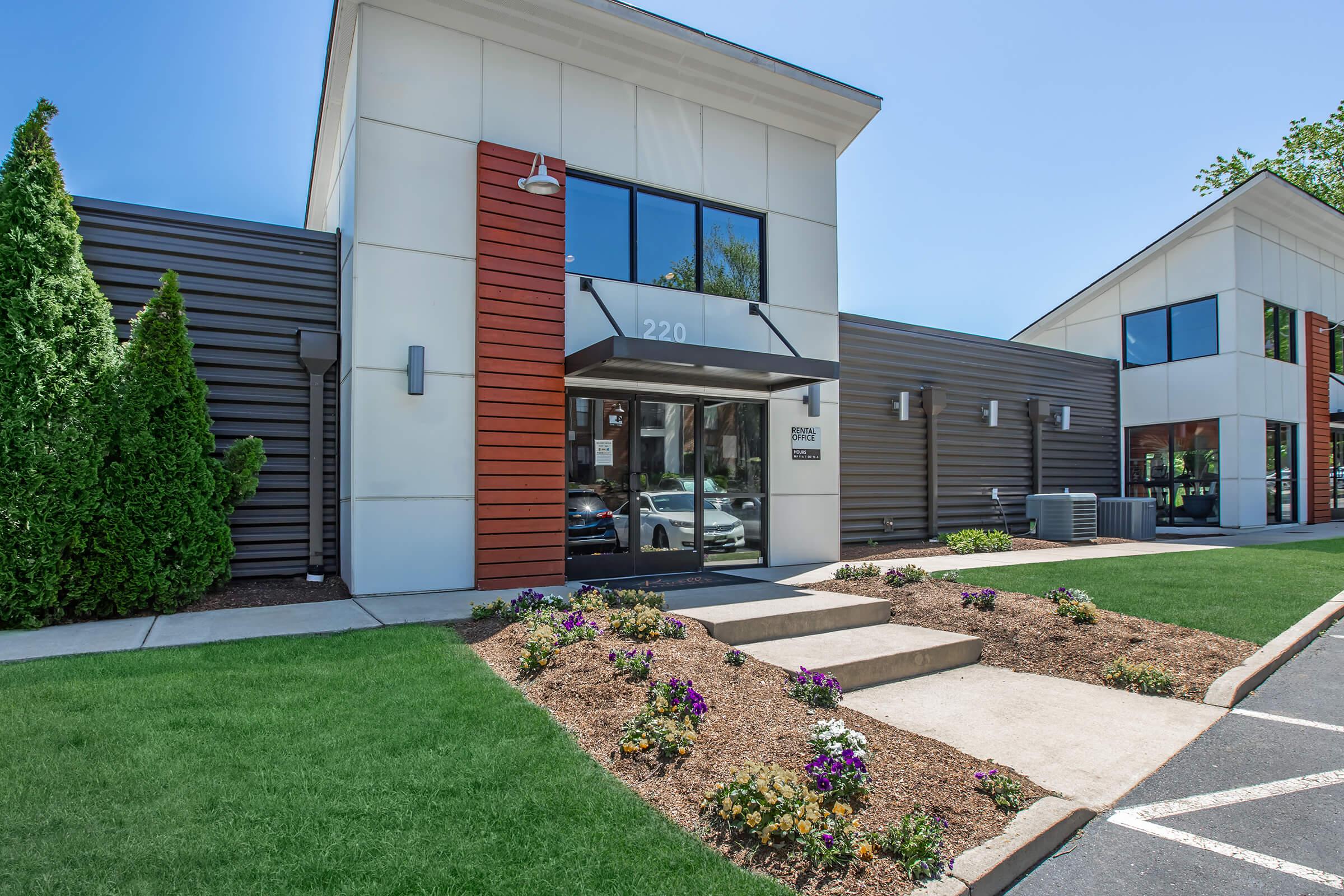 A modern building entrance featuring a sleek façade with wooden accents. Surrounding the entrance are landscaped flower beds with colorful plants, and a well-maintained lawn. Clear skies and bright sunlight enhance the contemporary architecture of the structure.