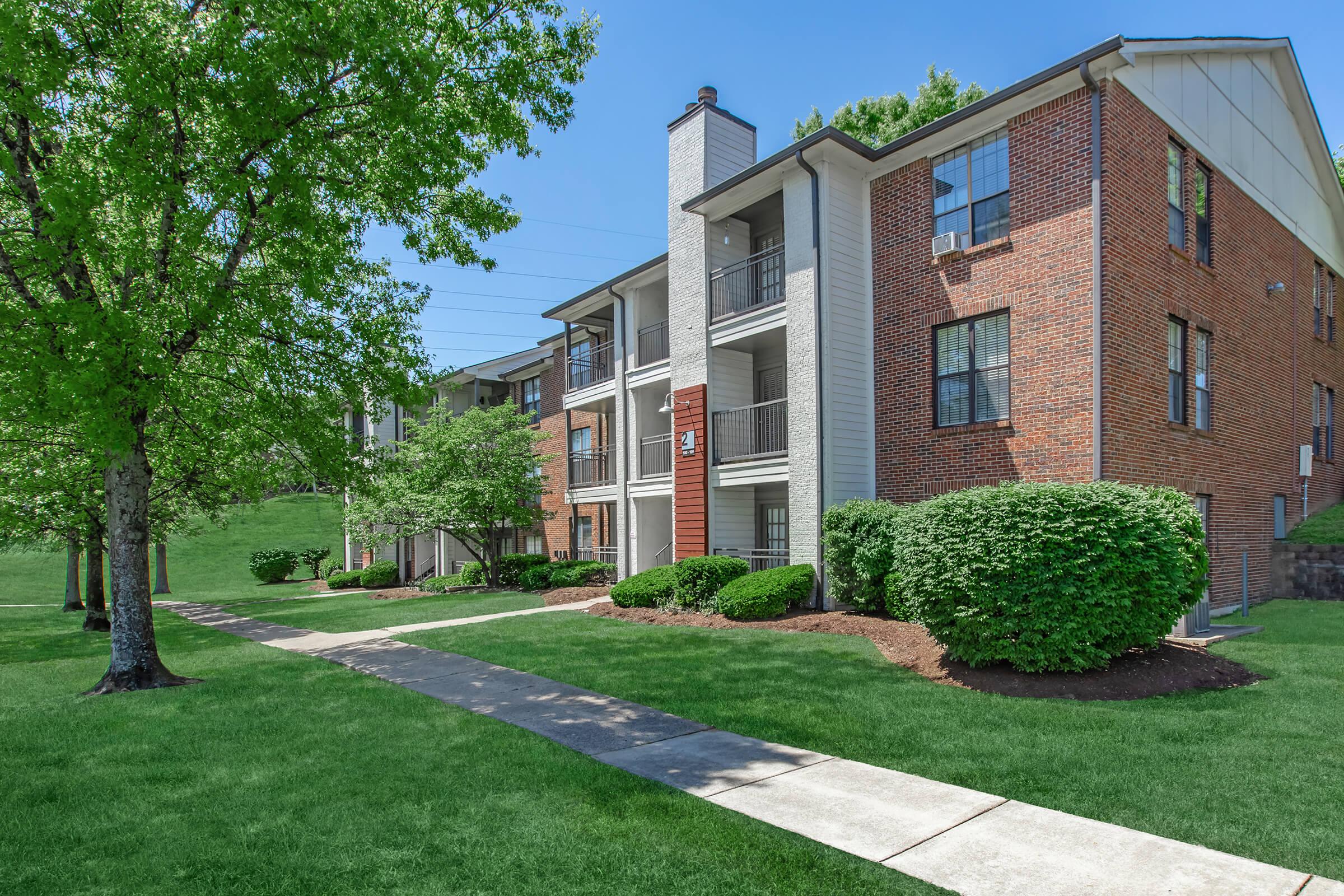 A well-maintained apartment complex with brick exterior and balconies, surrounded by lush green grass, trees, and shrubs. A concrete walkway runs through the landscaped area, leading to the building entrances under a clear blue sky.