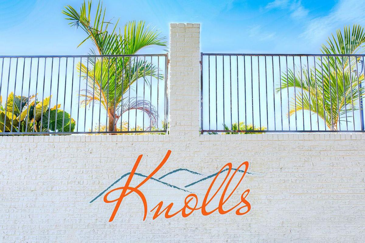A wall with the word "Knolls" in a stylized orange font. Above the wall, there are green palm trees and a clear blue sky, along with a black metal fence. The scene conveys a tropical or coastal ambiance.