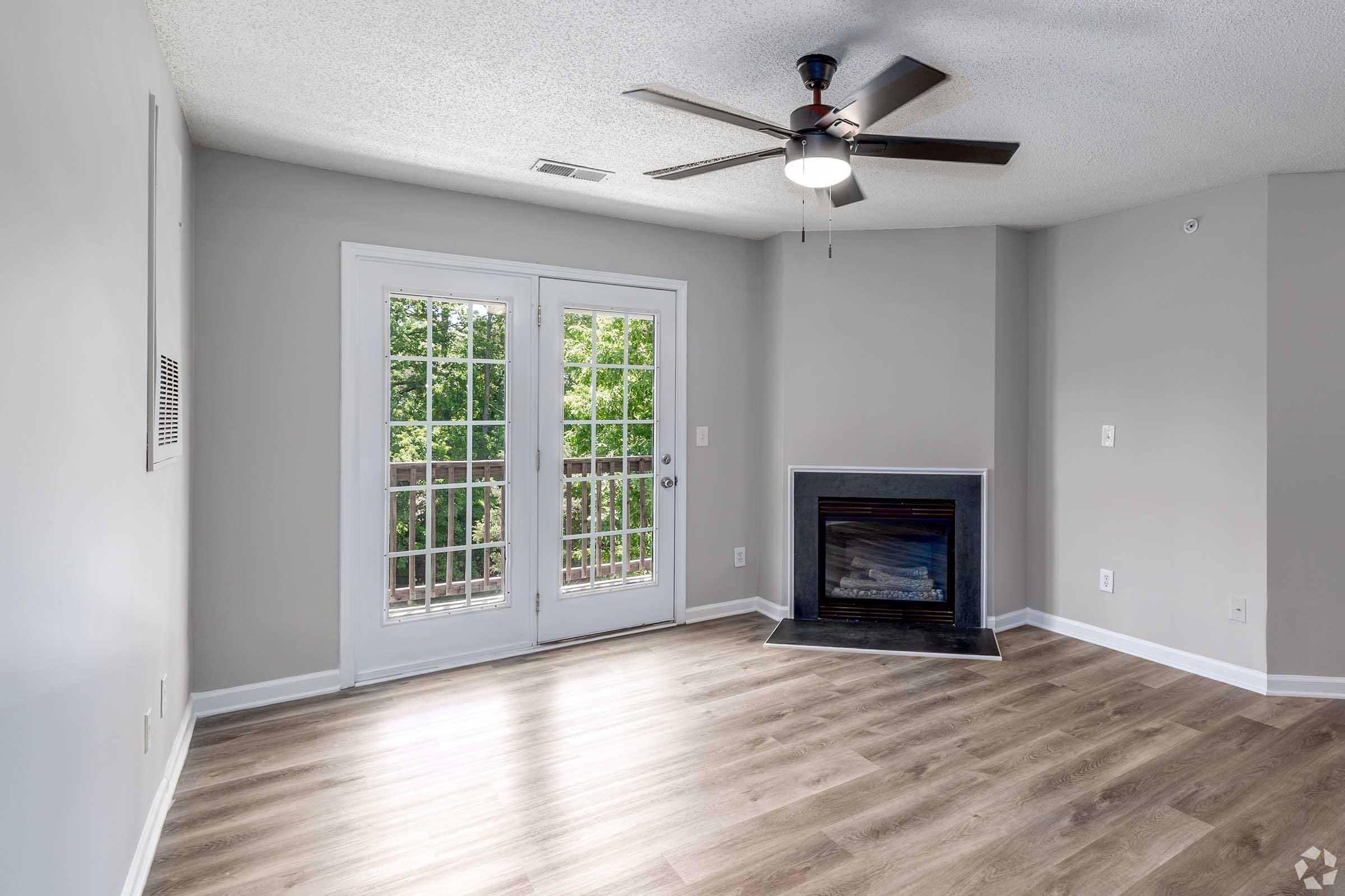 A spacious, light-filled living room with a ceiling fan, hardwood-style flooring, and a modern electric fireplace. Two glass doors lead to a balcony, while the walls are painted in neutral tones, creating an inviting atmosphere.