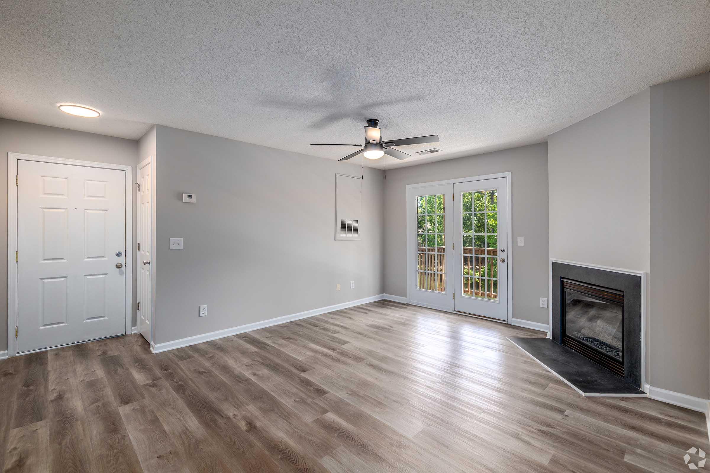 Spacious living area featuring light gray walls, a ceiling fan, and light wood flooring. To the right, there is a large window with sliding glass doors that lead to an outdoor space. A modern fireplace is situated in the corner, adding warmth to the room. A front door is visible on the left.