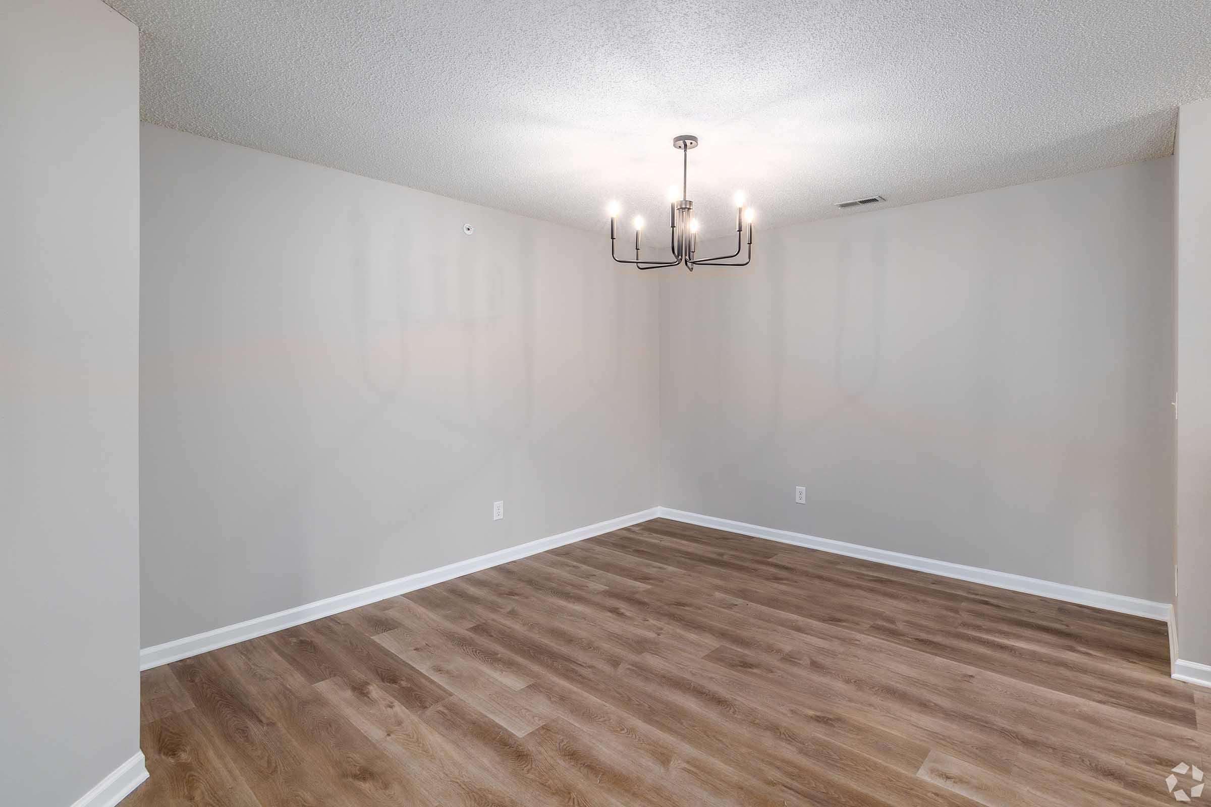 An empty room with light gray walls and a wood laminate floor. A modern chandelier hangs from the ceiling, casting shadows on the wall. The space is bright and uncluttered, suitable for various uses such as a dining area or living space.